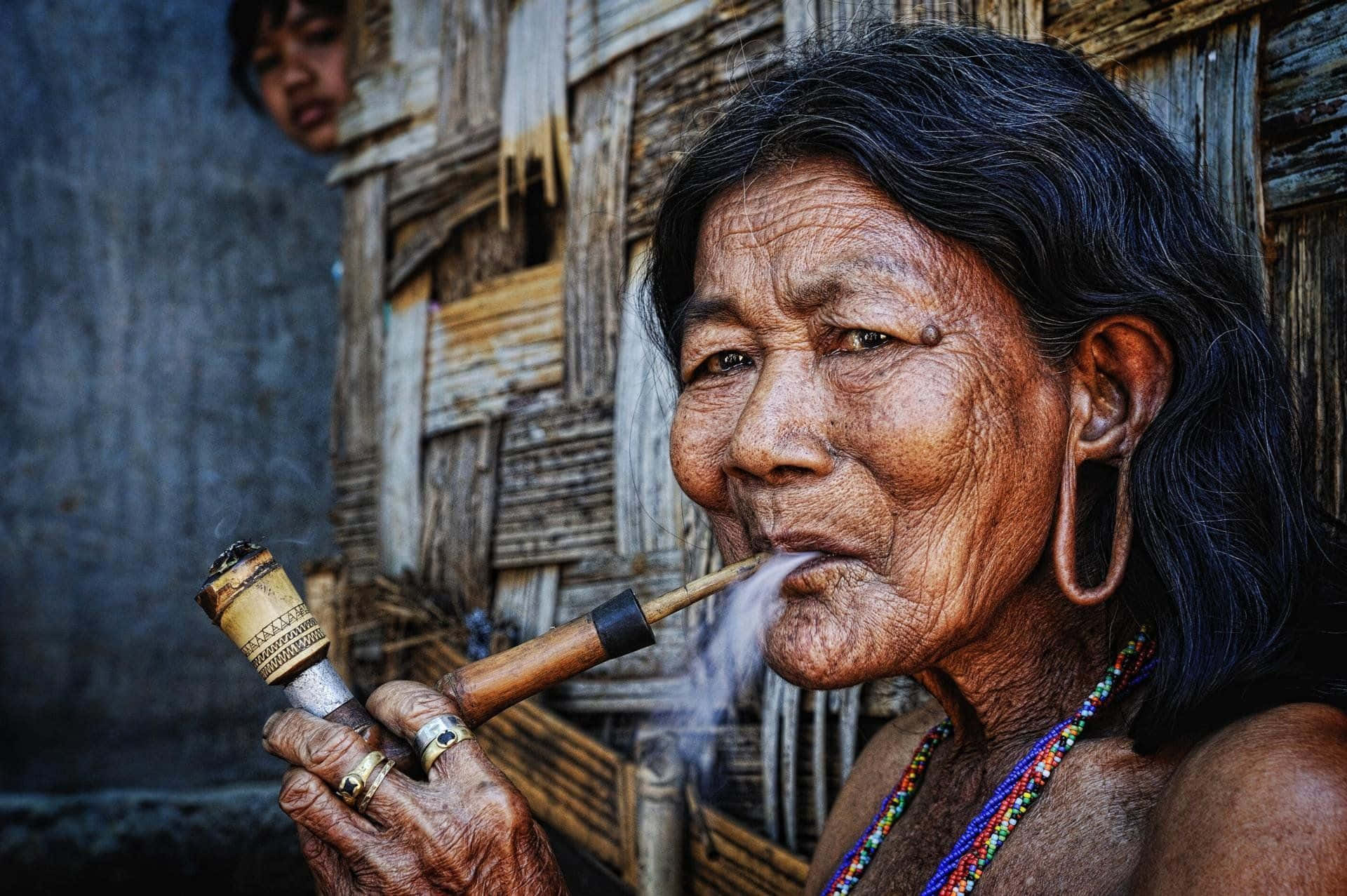 Old Lady 桌布 壁紙 — 一位滿臉皺紋、黑髮的老婦人，戴著色彩繽紛的珠鏈，在風化的木屋背景下抽著木煙斗。