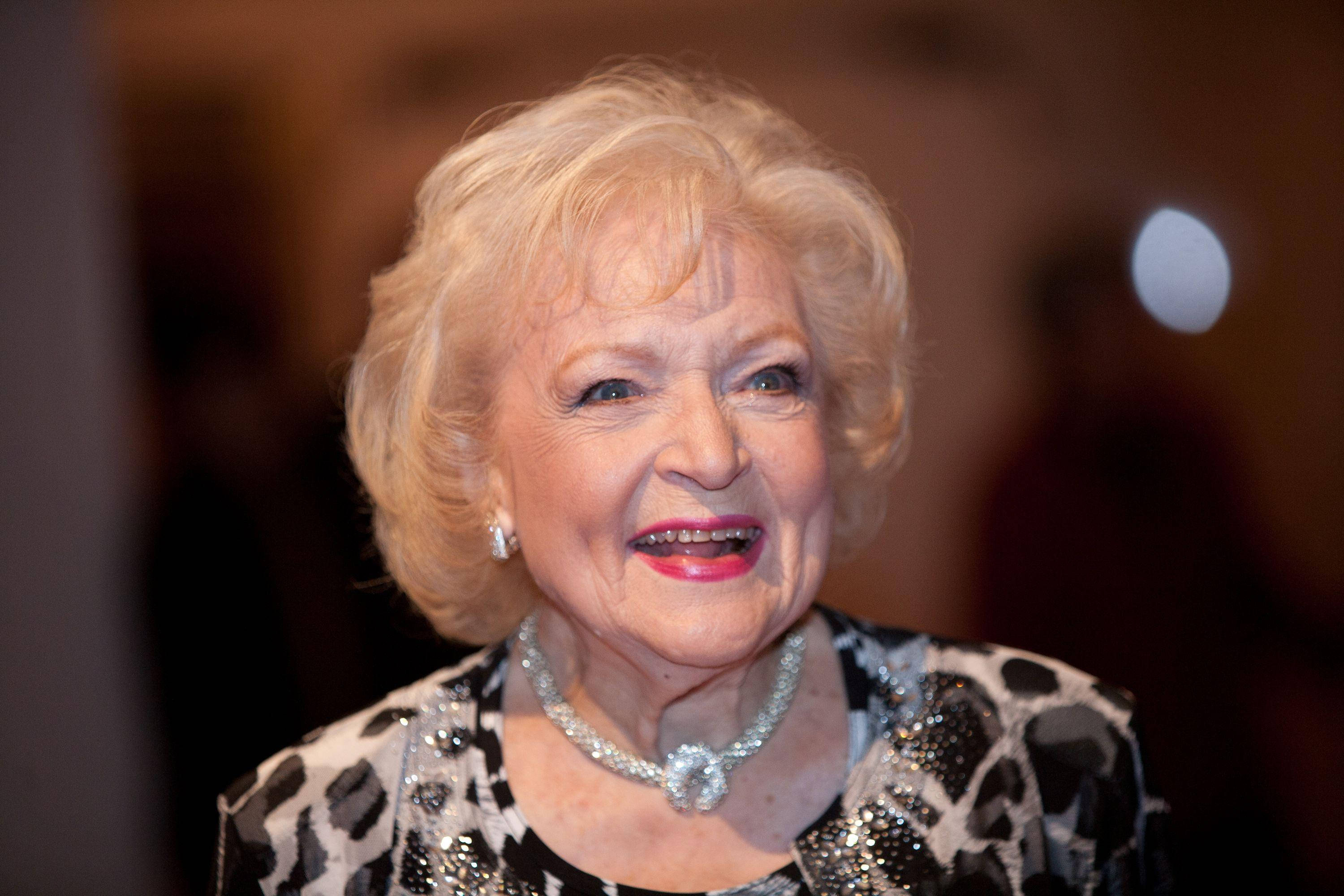 Betty White 桌布 壁紙 — 一位滿面紅光的銀髮女士，金髮紅唇，穿著閃爍的黑白套裝，溫暖地微笑。