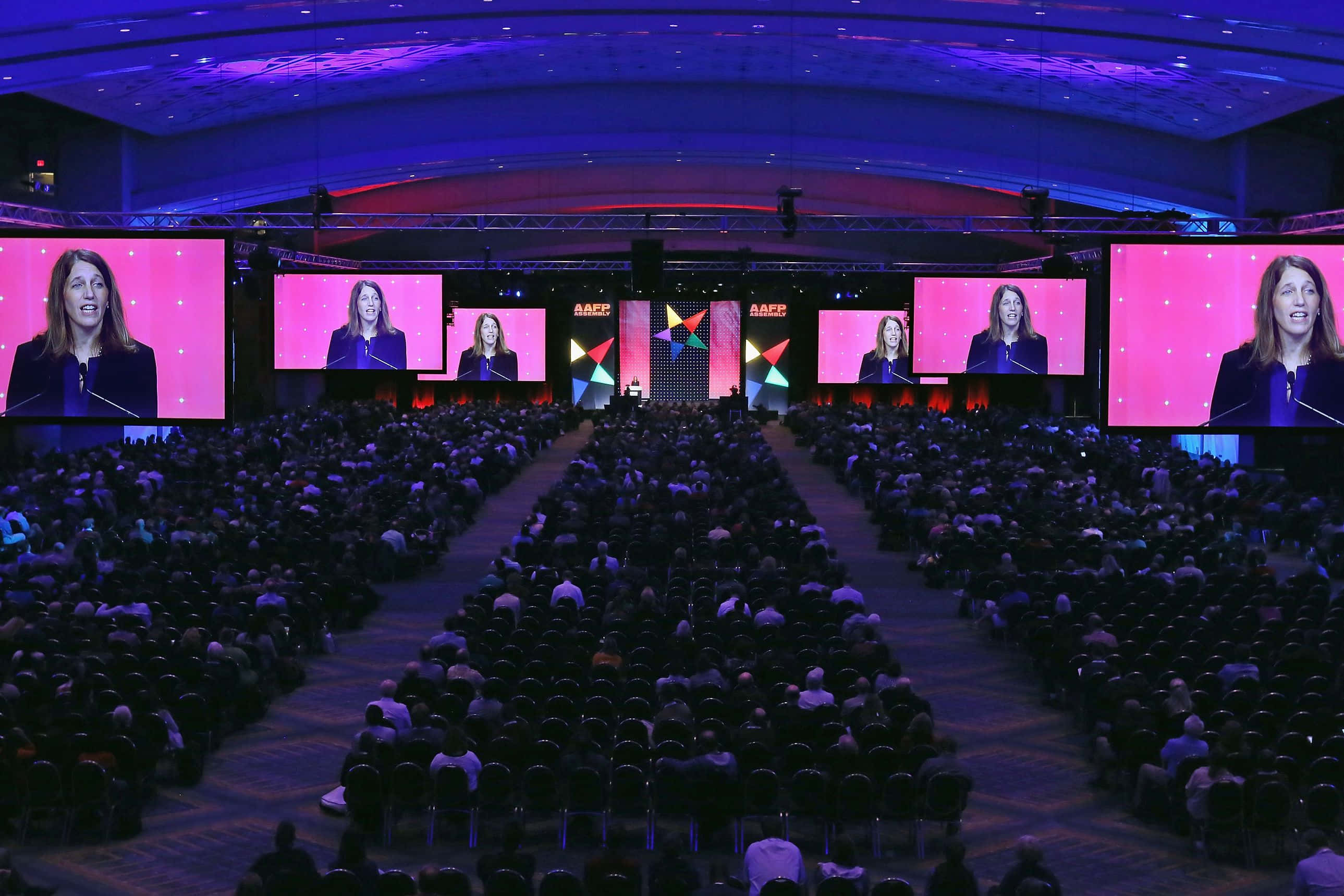 Corporate Event 桌布 壁紙 — 一位演講者在一個由充滿活力的紫色和粉紅色舞台燈光照亮的宏偉大廳中向大量觀眾發表演講。