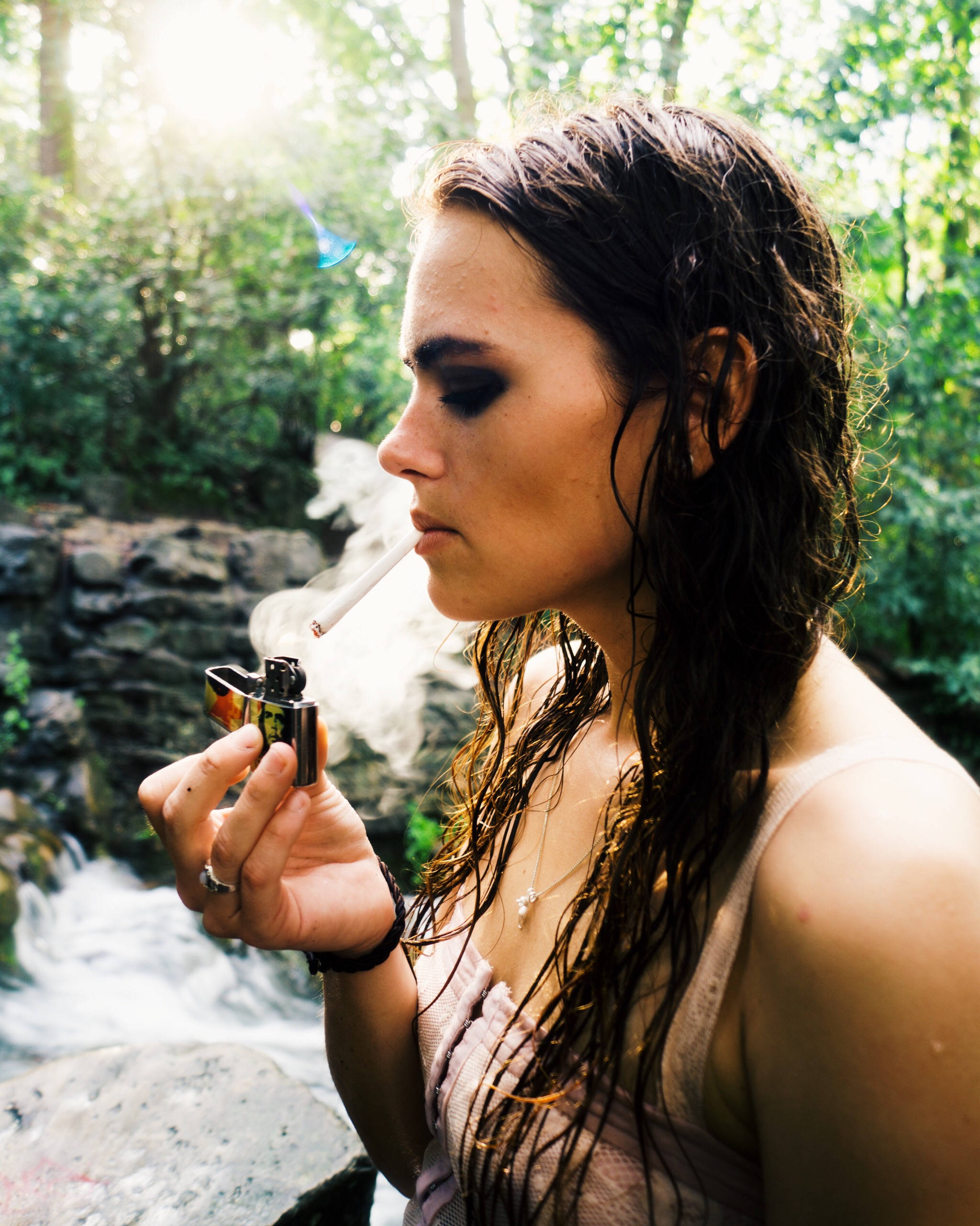 吸大麻 桌布 壁紙 — 一位濕髮女性在陽光明媚的森林溪流旁抽煙，手持一個色彩繽紛的打火機。
