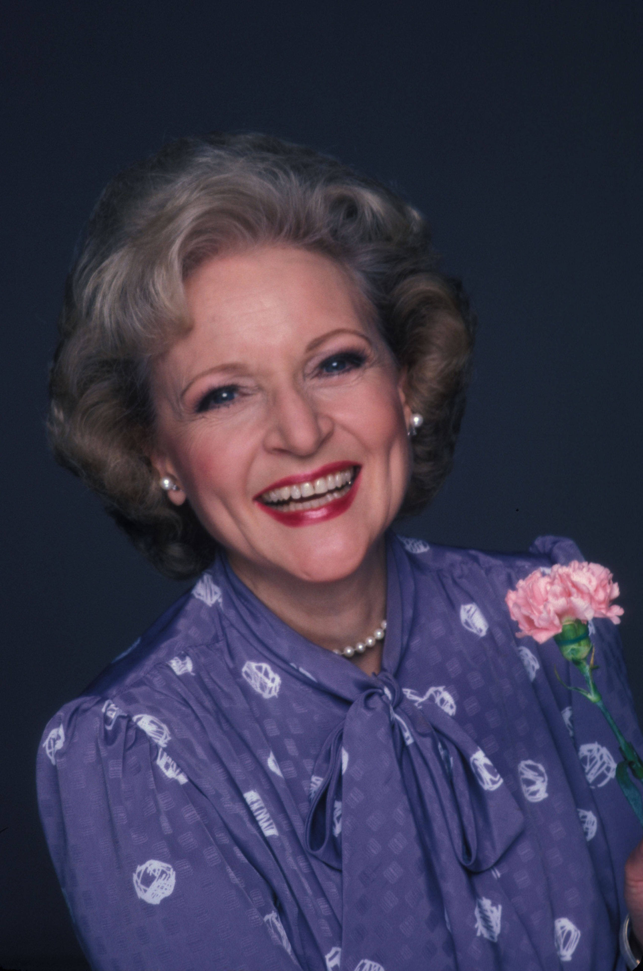 Betty White 桌布 壁紙 — 一位灰白捲髮的開朗老婦人，在深藍色背景前手持粉紅色康乃馨，露出燦爛的笑容。