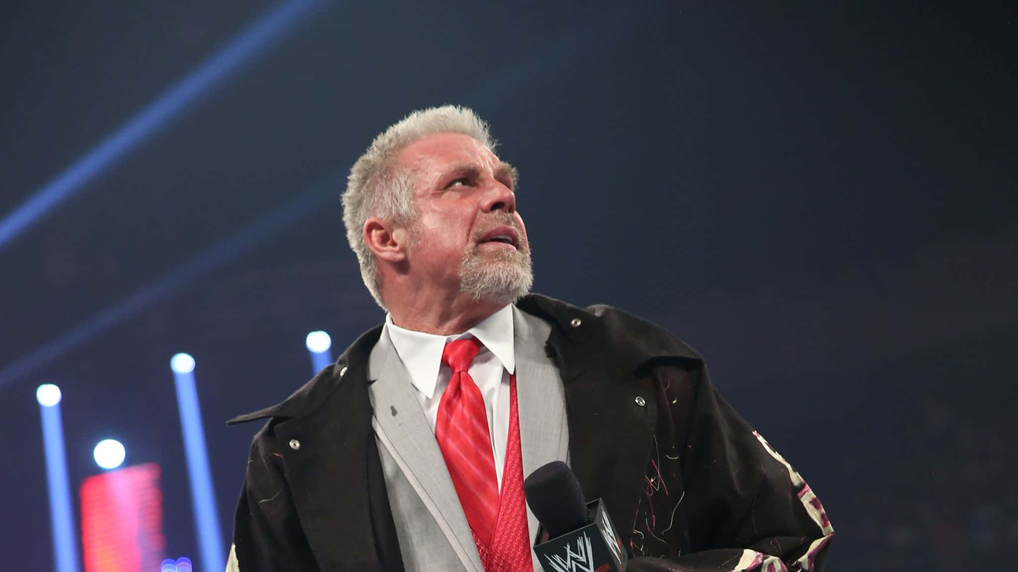 WWE 桌布 壁紙 — 一位灰白頭髮和鬍鬚的摔角手站在燈光璀璨的舞台上，身穿黑色外套、灰色西裝和紅色領帶，手持WWE麥克風。