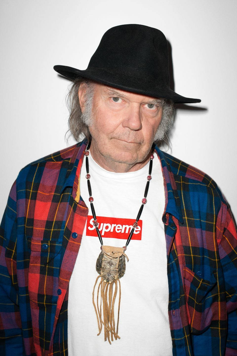 supreme 桌布 壁紙 — 一位灰白頭髮的年長男士戴著黑色軟呢帽、格紋襯衫和Supreme T恤，搭配珠鍊，背景為純白色。