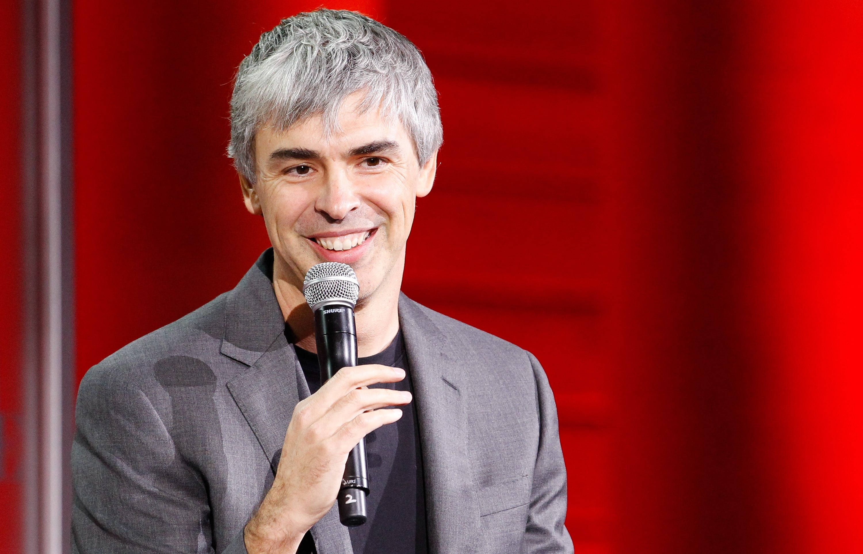 google 桌布 壁紙 — 一位灰白頭髮的微笑男子在鮮紅色背景下拿著麥克風。