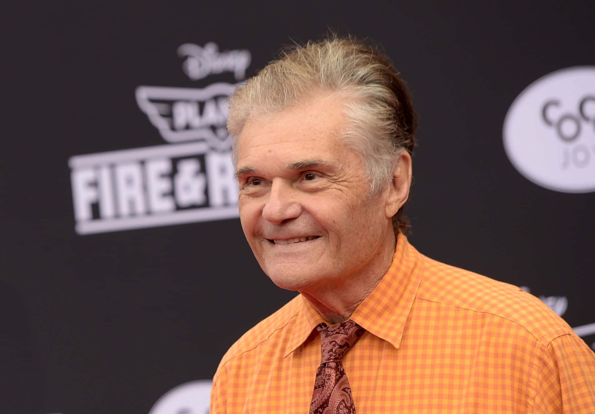 Fred Willard 桌布 壁紙 — 一位灰白頭髮的微笑長者，在迪士尼活動中穿著橙色格紋襯衫和圖案領帶。