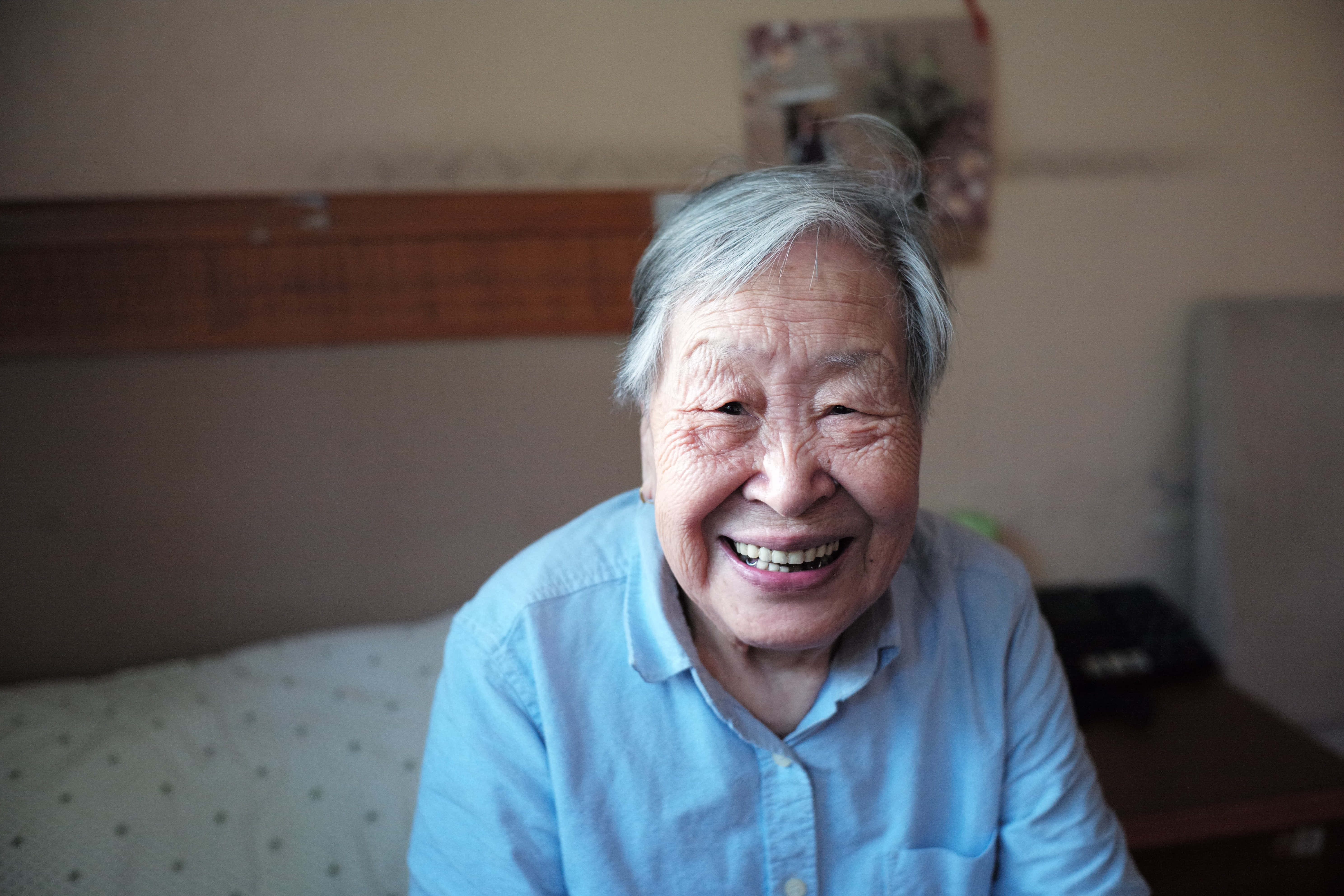 smiling 桌布 壁紙 — 一位灰白頭髮的老年女性在柔和光線的臥室中溫暖地微笑，身穿淺藍色襯衫。
