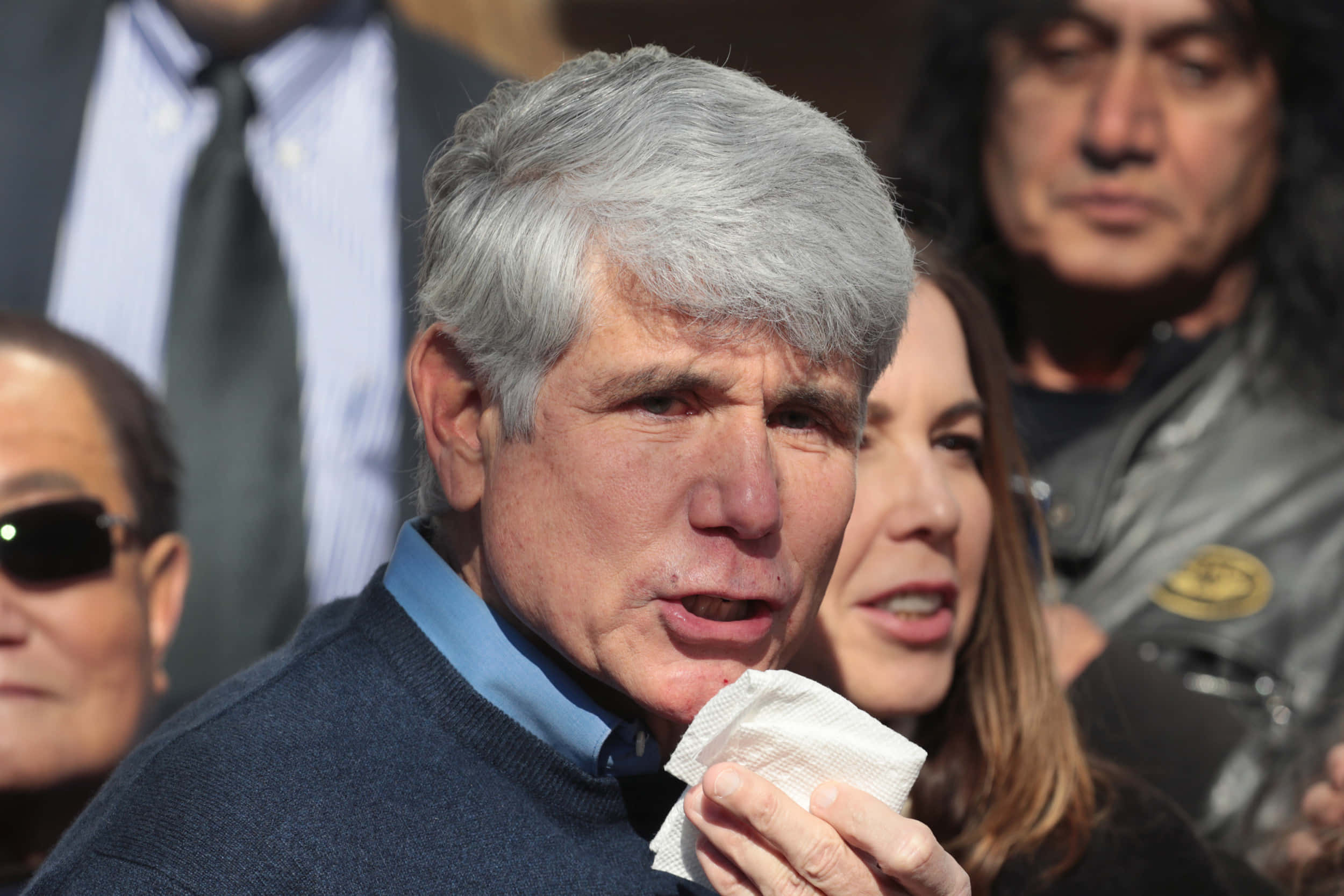 政治 桌布 壁紙 — 一位灰白頭髮的長者，在明亮的戶外環境中被人群包圍，手持紙巾靠近嘴巴。