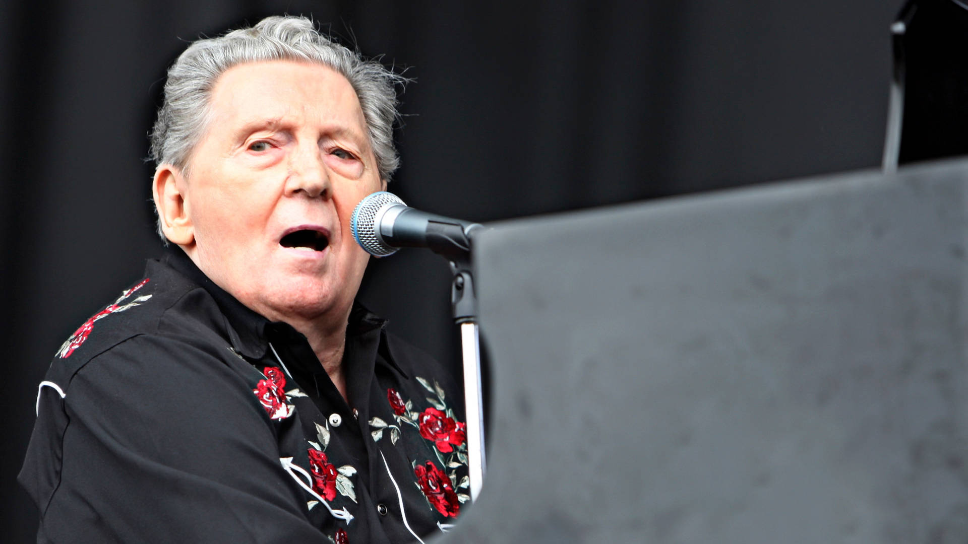 Jerry Lee Lewis 桌布 壁紙 — 一位灰白頭髮的長者坐在舞台上的鋼琴前，熱情地對著麥克風唱歌。