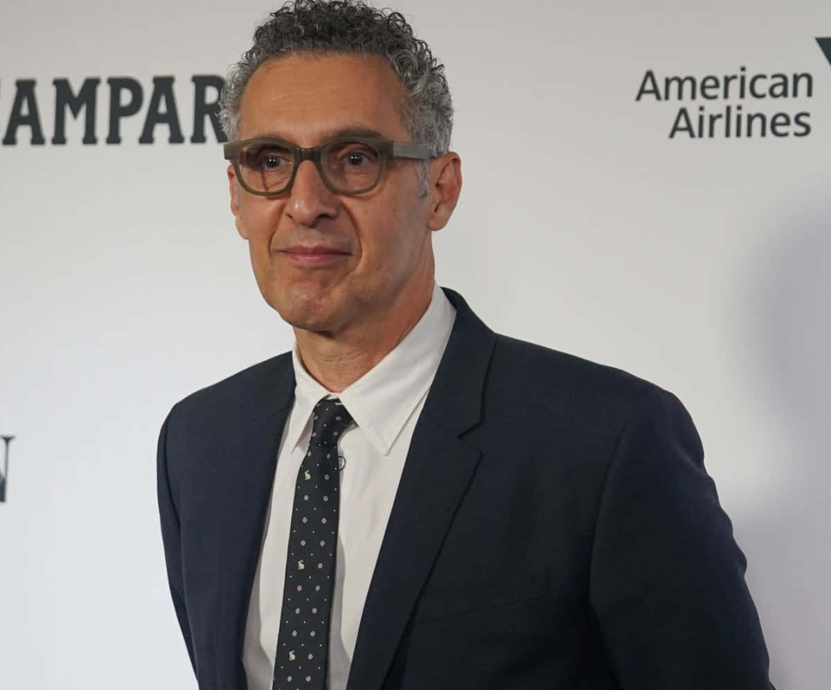 John Turturro 桌布 壁紙 — 一位灰色捲髮戴眼鏡的男子，身穿深色西裝和圓點領帶，背景是品牌活動場景。