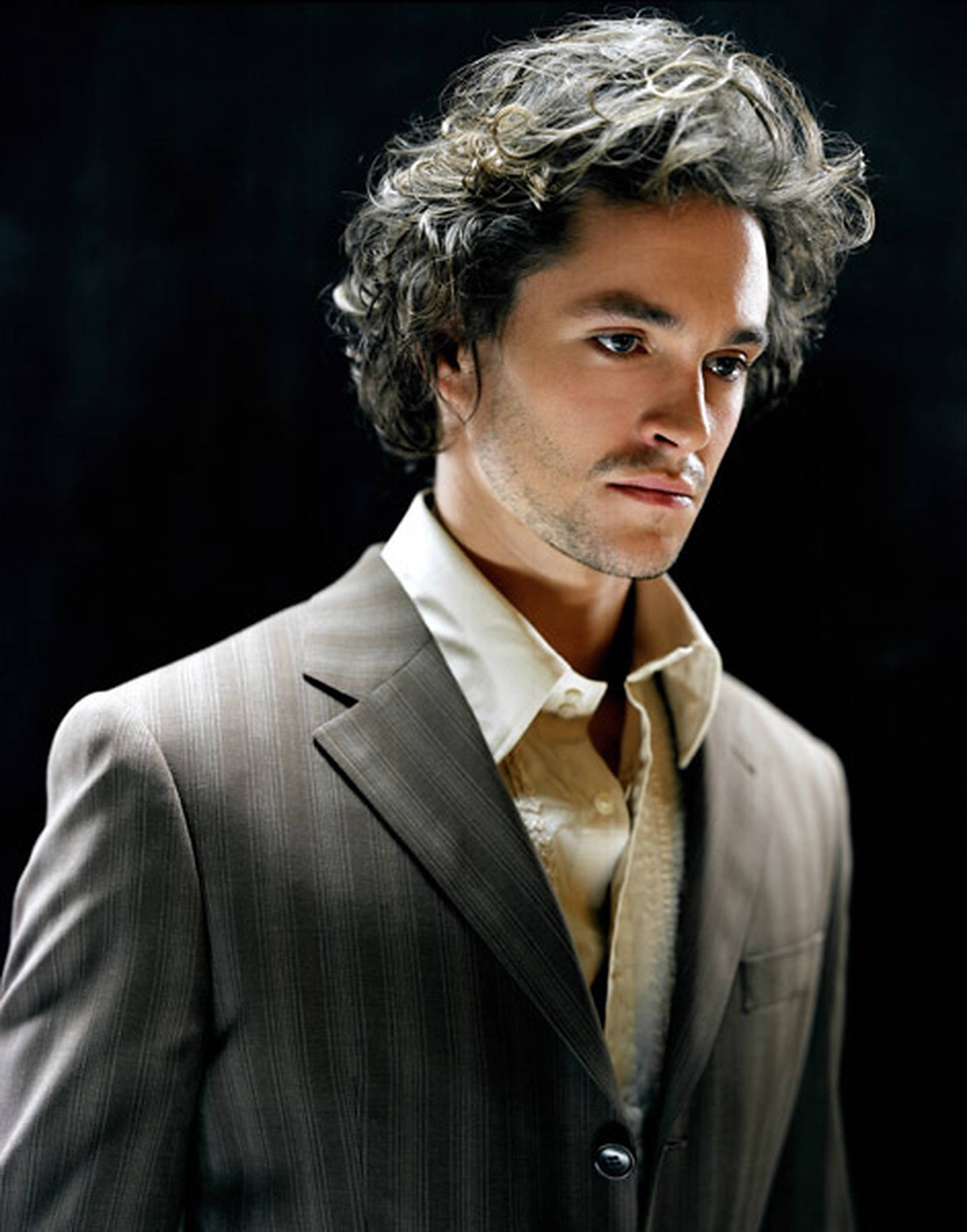 Hugh Dancy 桌布 壁紙 — 一位灰色捲髮男士穿著細條紋西裝，背景深色，光線柔和。