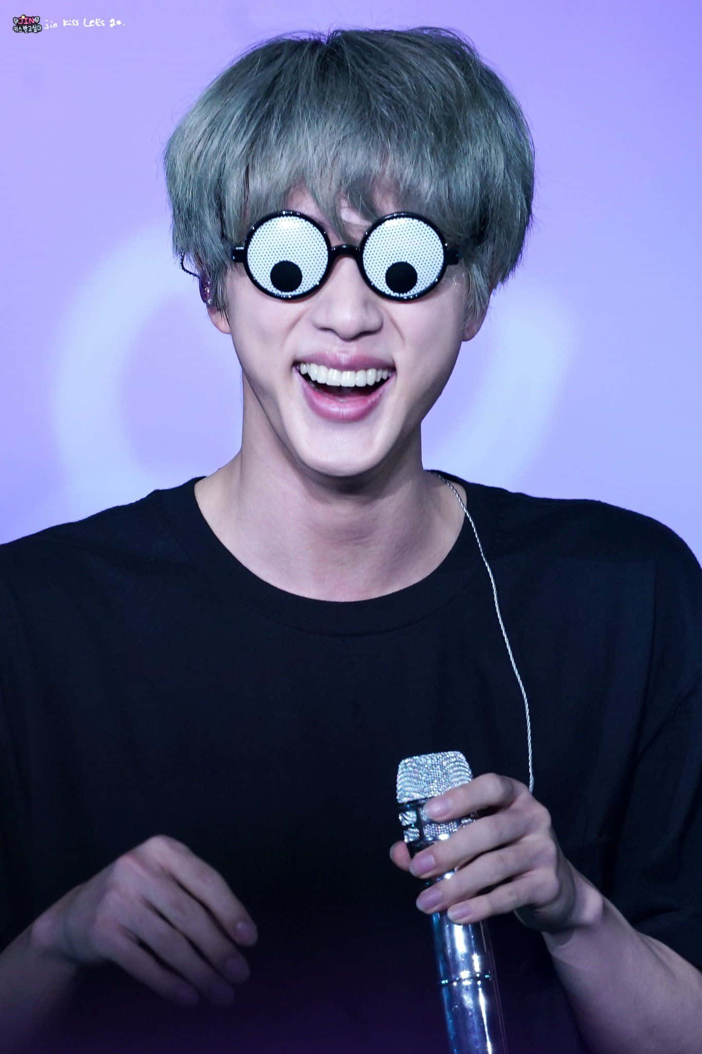 BTS 桌布 壁紙 — 一位灰髮、戴著超大凸眼眼鏡的開朗表演者，在紫色舞台燈光下微笑著手持閃亮麥克風。