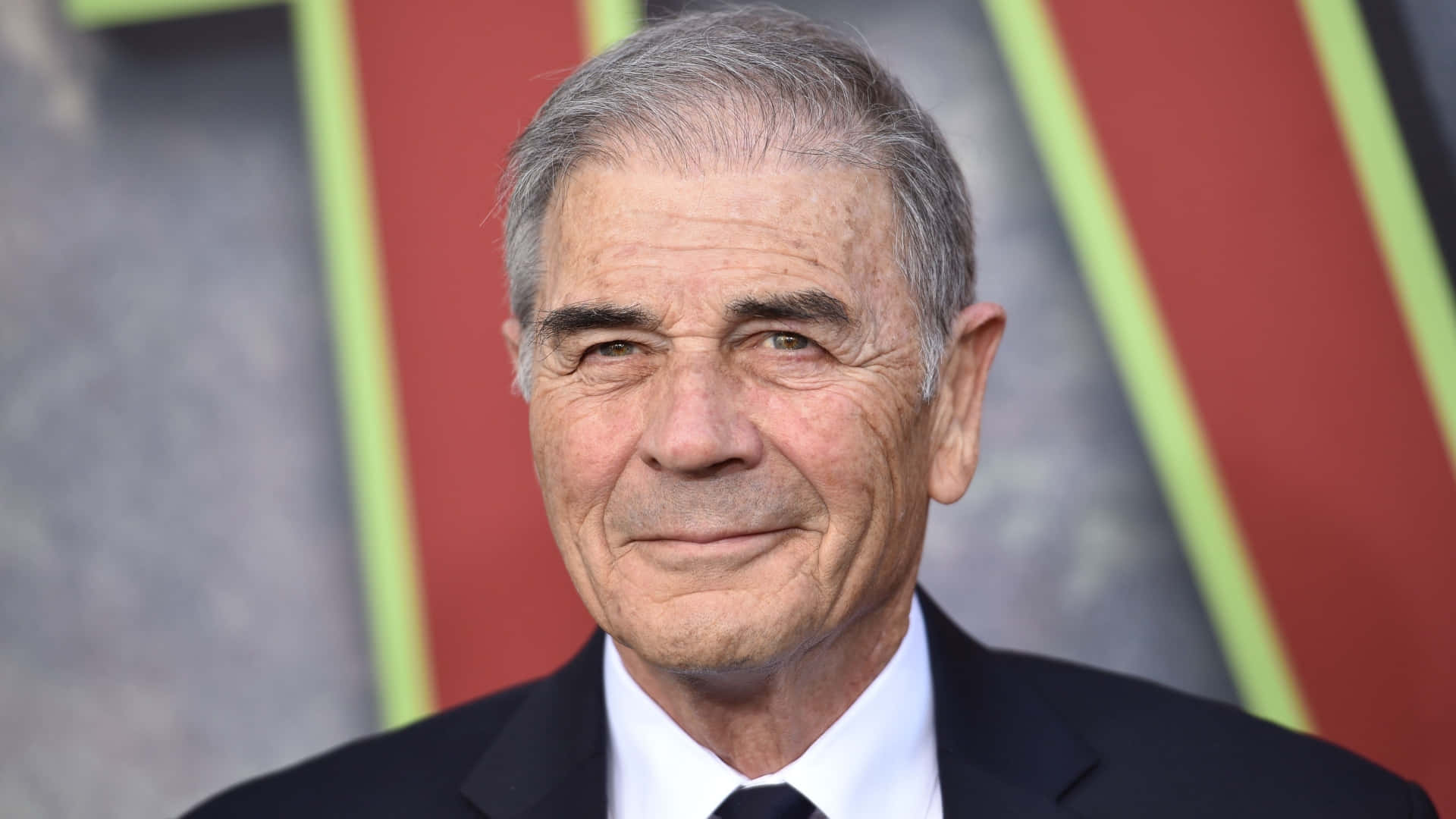 actor 桌布 壁紙 — 一位灰髮、穿著深色西裝和領帶的傑出老年男士，在色彩繽紛的幾何背景下的特寫肖像。