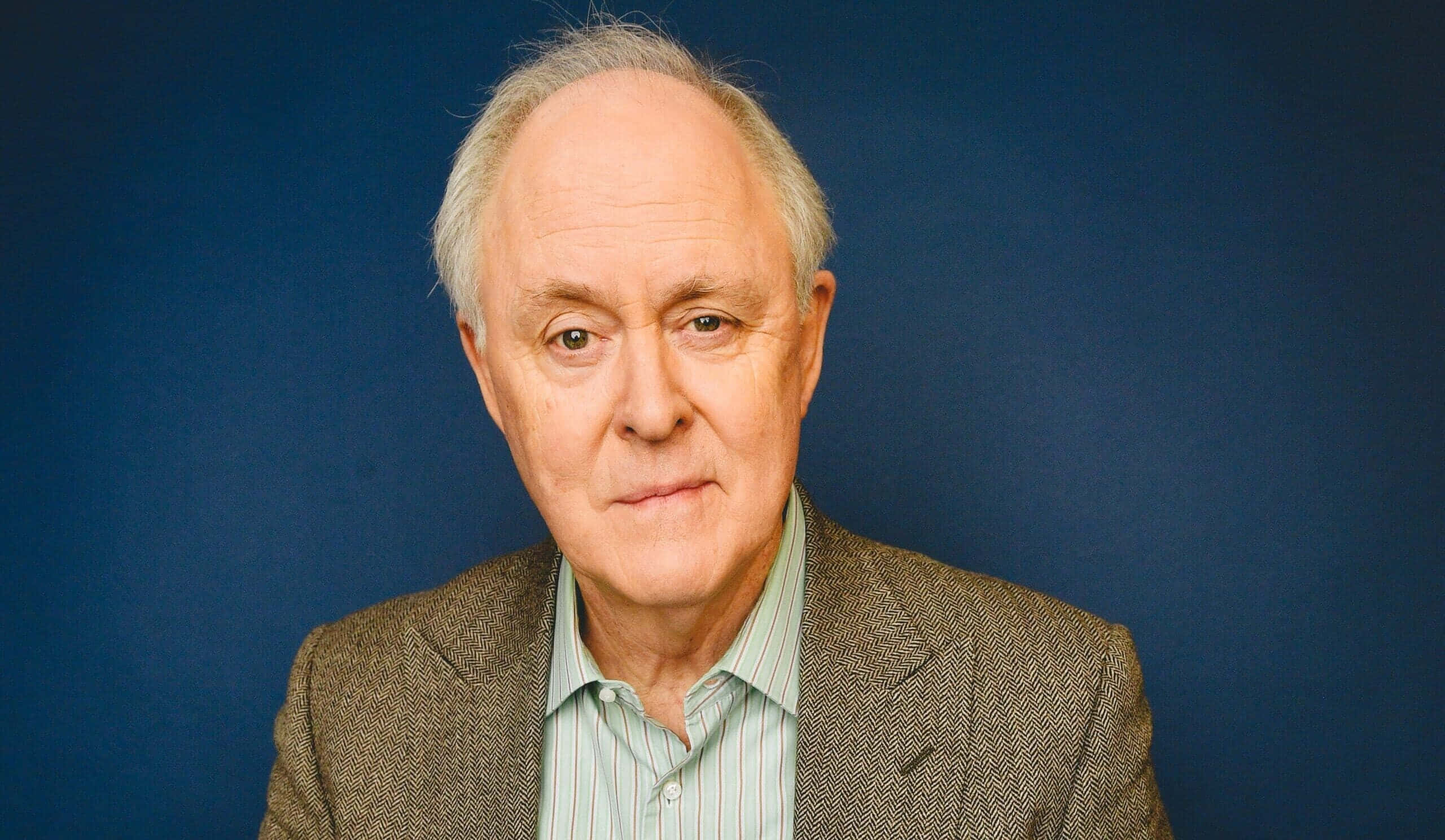 John Lithgow 桌布 壁紙 — 一位灰髮、表情嚴肅的老年男子，身穿棕色粗花呢外套，內搭條紋襯衫，背景為純藍色。