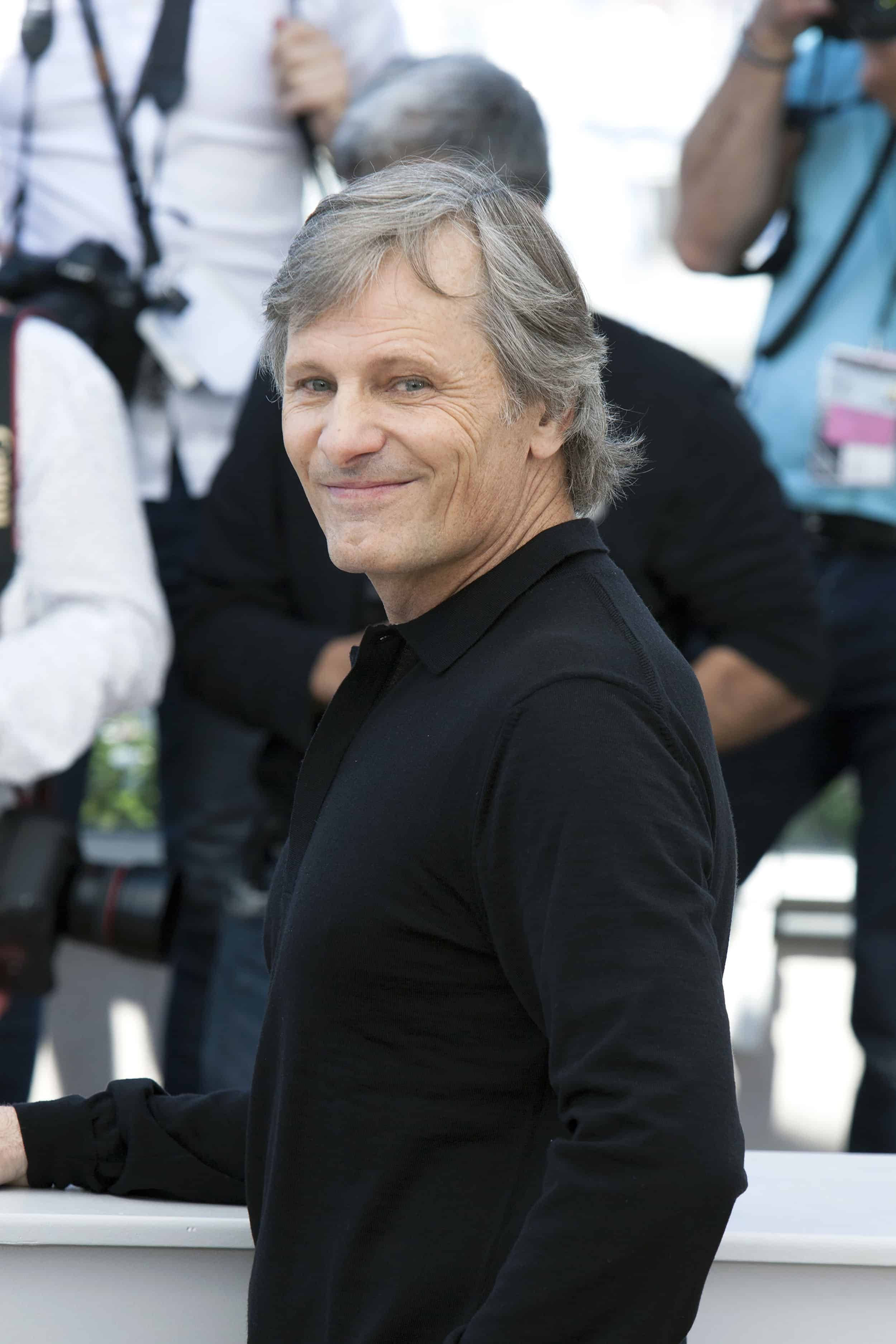actor 桌布 壁紙 — 一位灰髮中年男子在公共活動中靠在白色欄杆上，背景有攝影師，露出溫暖的笑容。