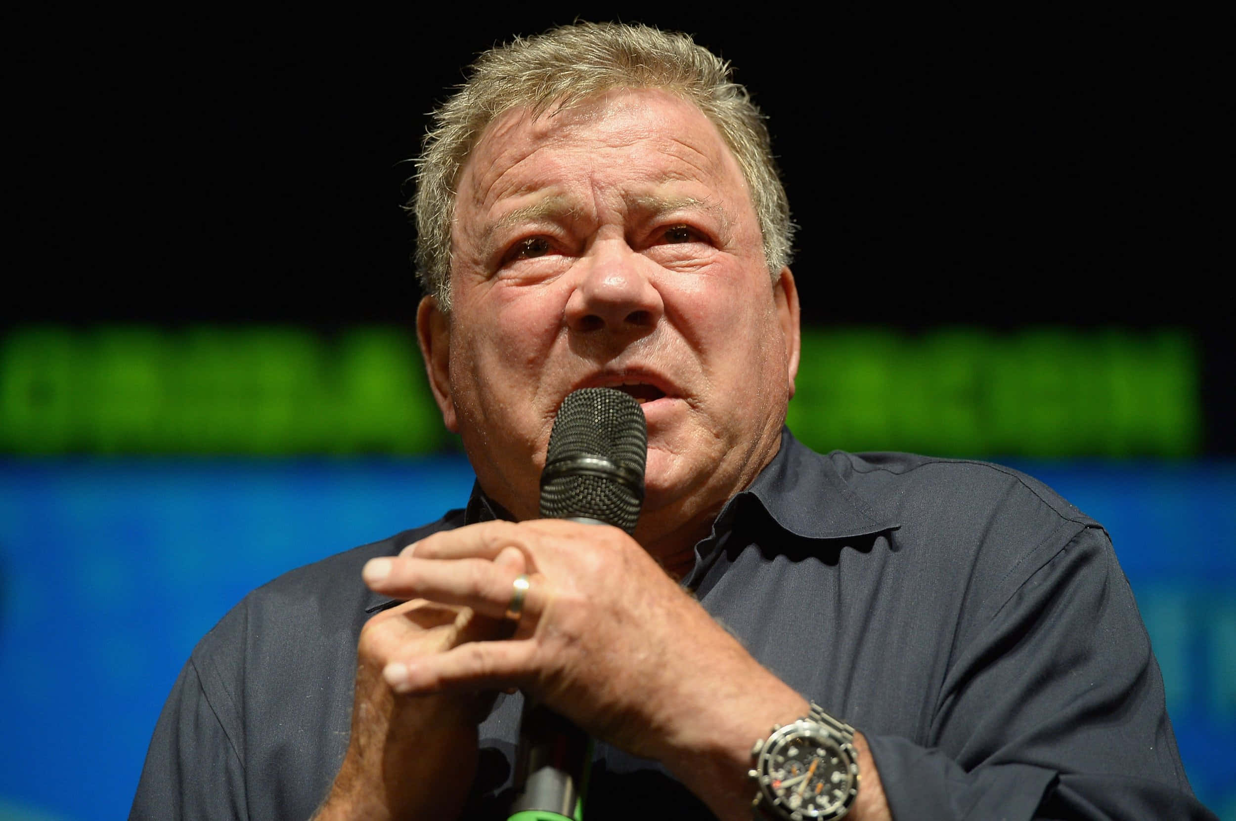 william shatner 桌布 壁紙 — 一位灰髮中年男子在綠色和藍色背景燈光的座談會上對著麥克風發言。