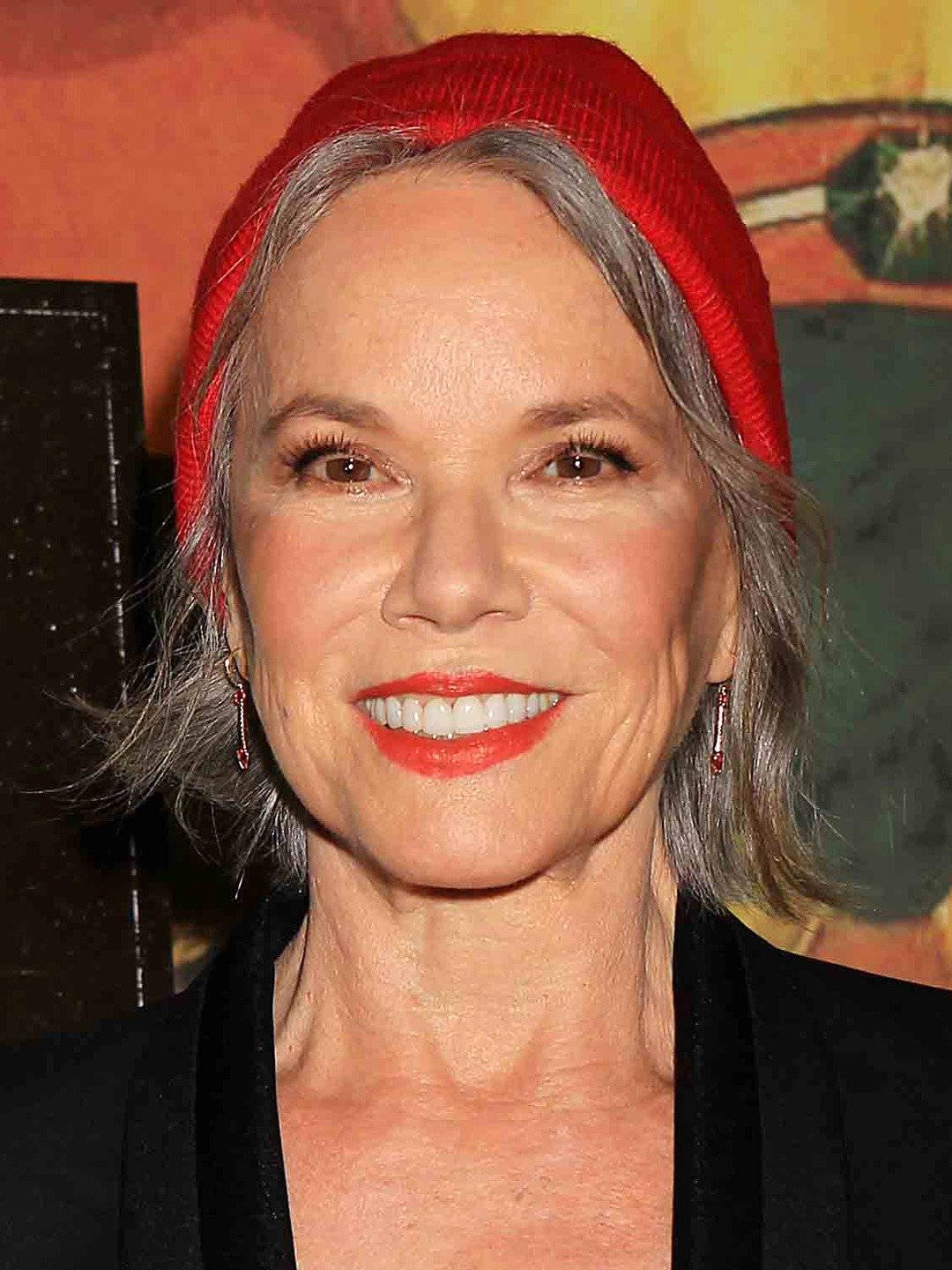 Celebrity 桌布 壁紙 — 一位灰髮微笑的女性戴著紅色毛帽和鮮紅色唇膏，背景是色彩繽紛的抽象圖案。