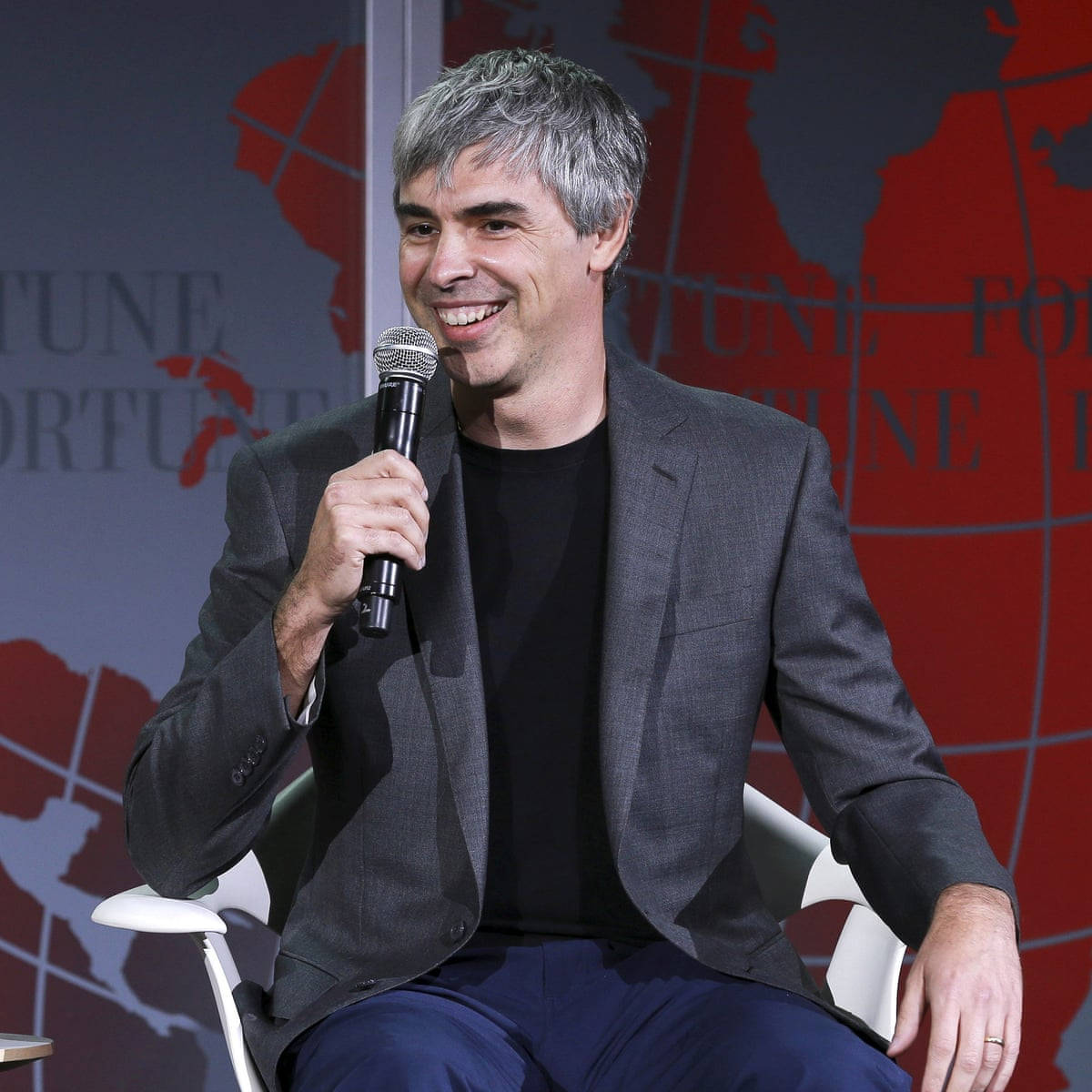 google 桌布 壁紙 — 一位灰髮微笑的男子坐在白色椅子上，手持麥克風，背景為紅色和灰色的Fortune品牌背景。