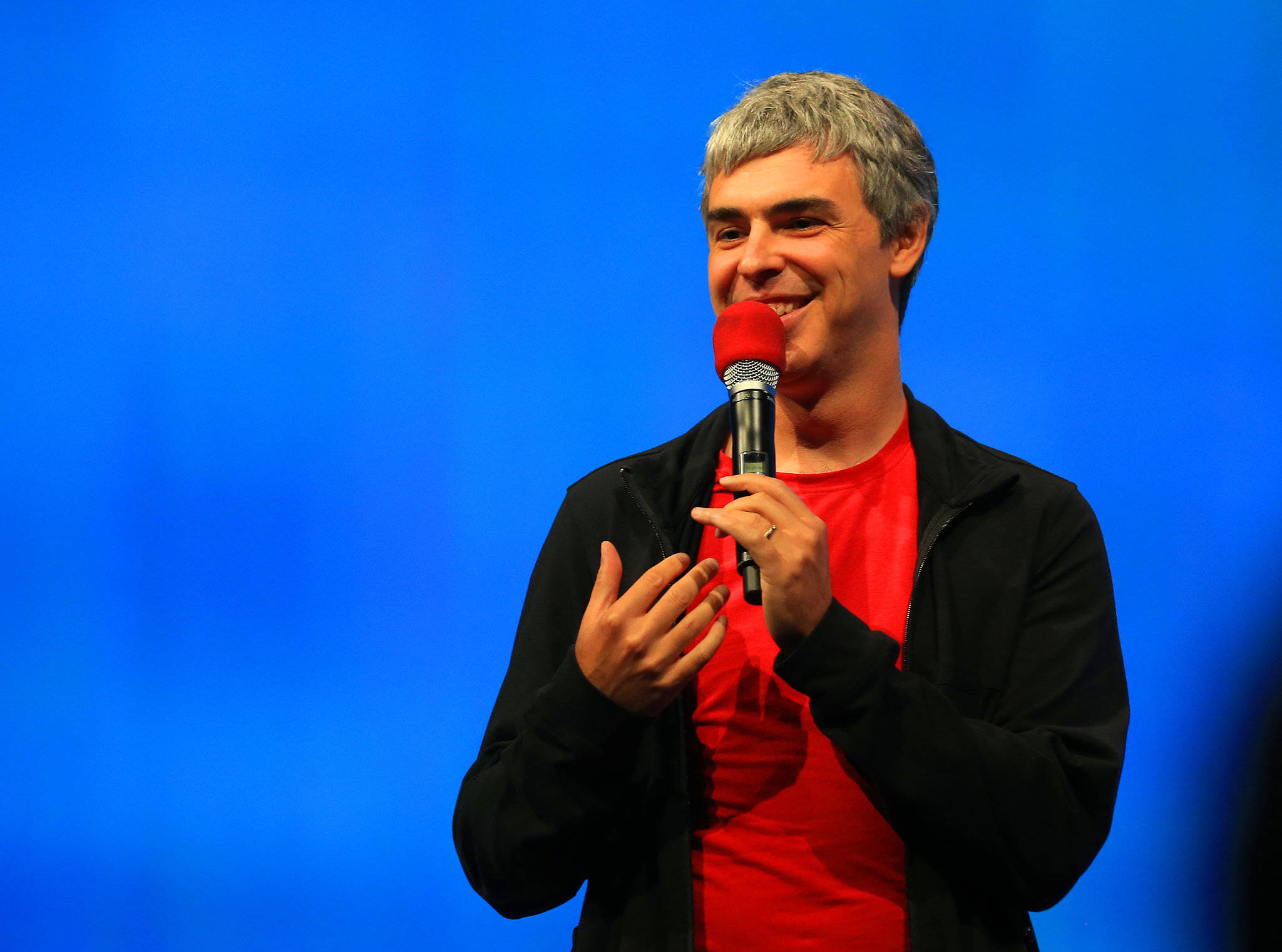 google 桌布 壁紙 — 一位灰髮微笑的男子手持紅色麥克風，背景為鮮豔的藍色，另一隻手做著手勢。
