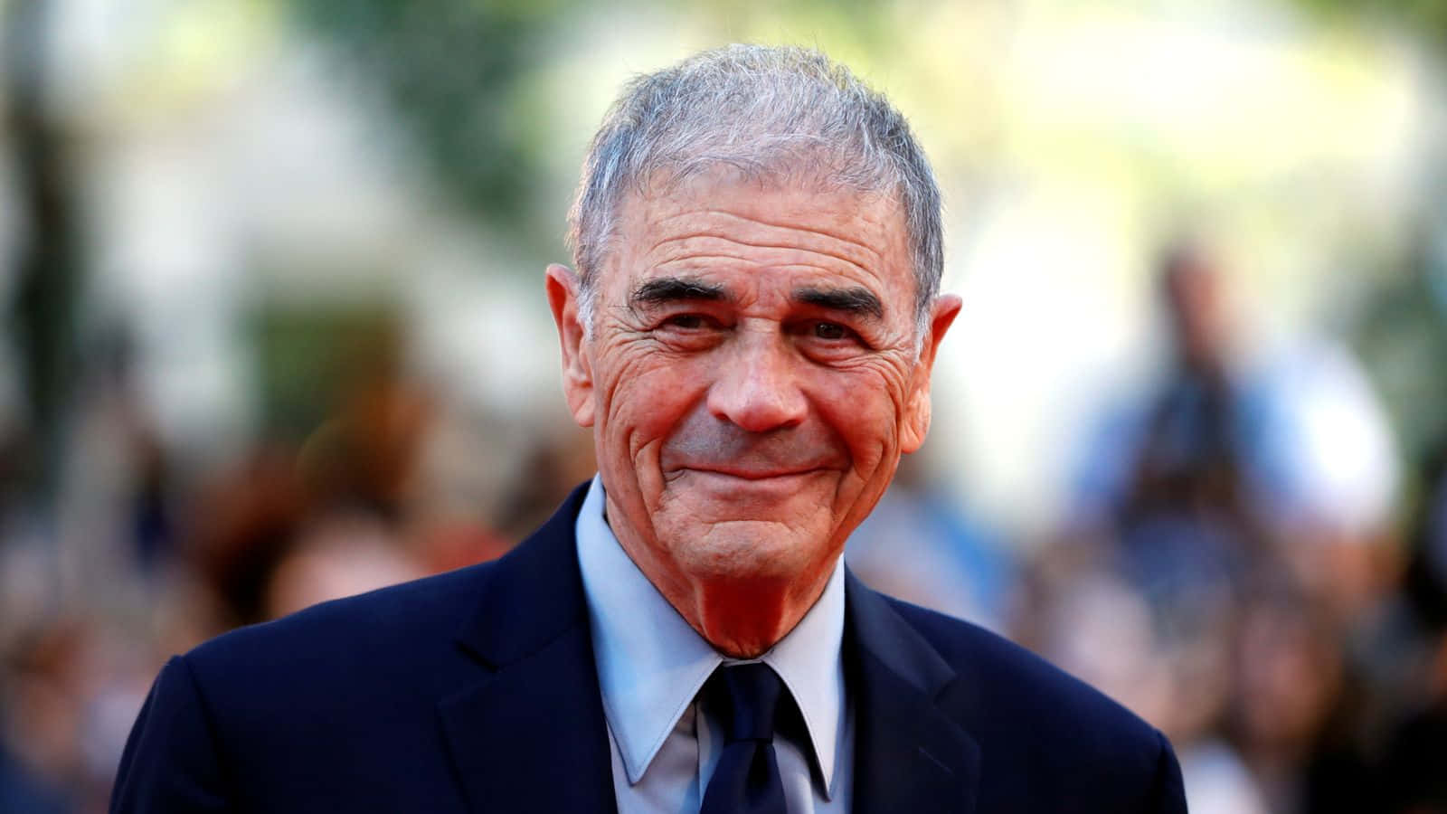 actor 桌布 壁紙 — 一位灰髮微笑的老年男子穿著深色西裝和領帶，背景是模糊的戶外環境的特寫肖像。
