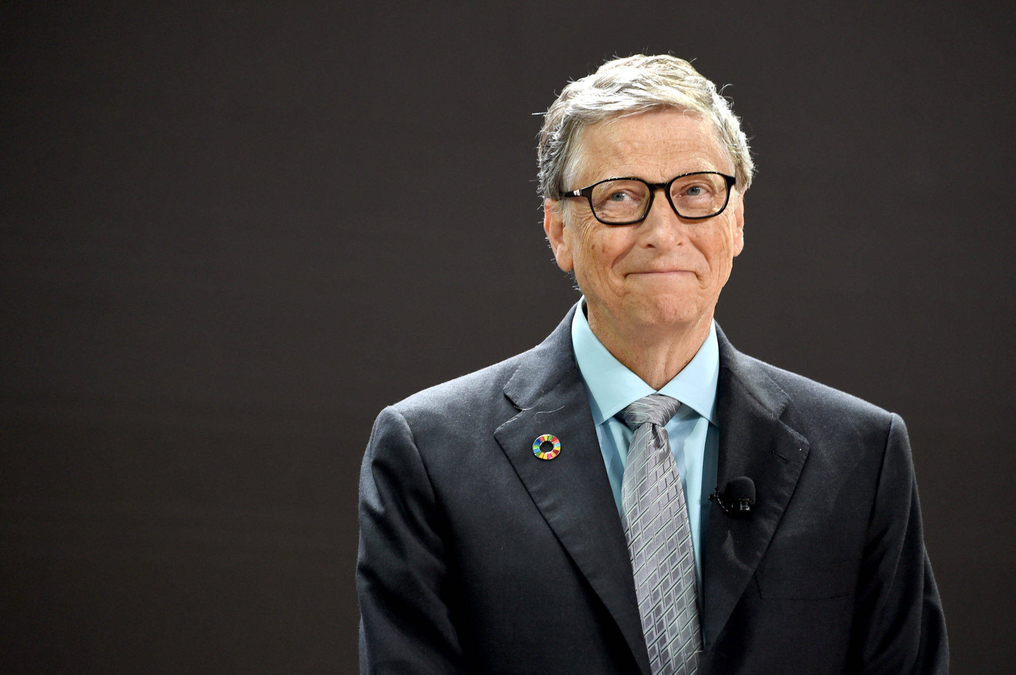 gates 桌布 壁紙 — 一位灰髮戴眼鏡的男士，身穿深色西裝、淺藍色襯衫和圖案領帶，背景為純色深色。