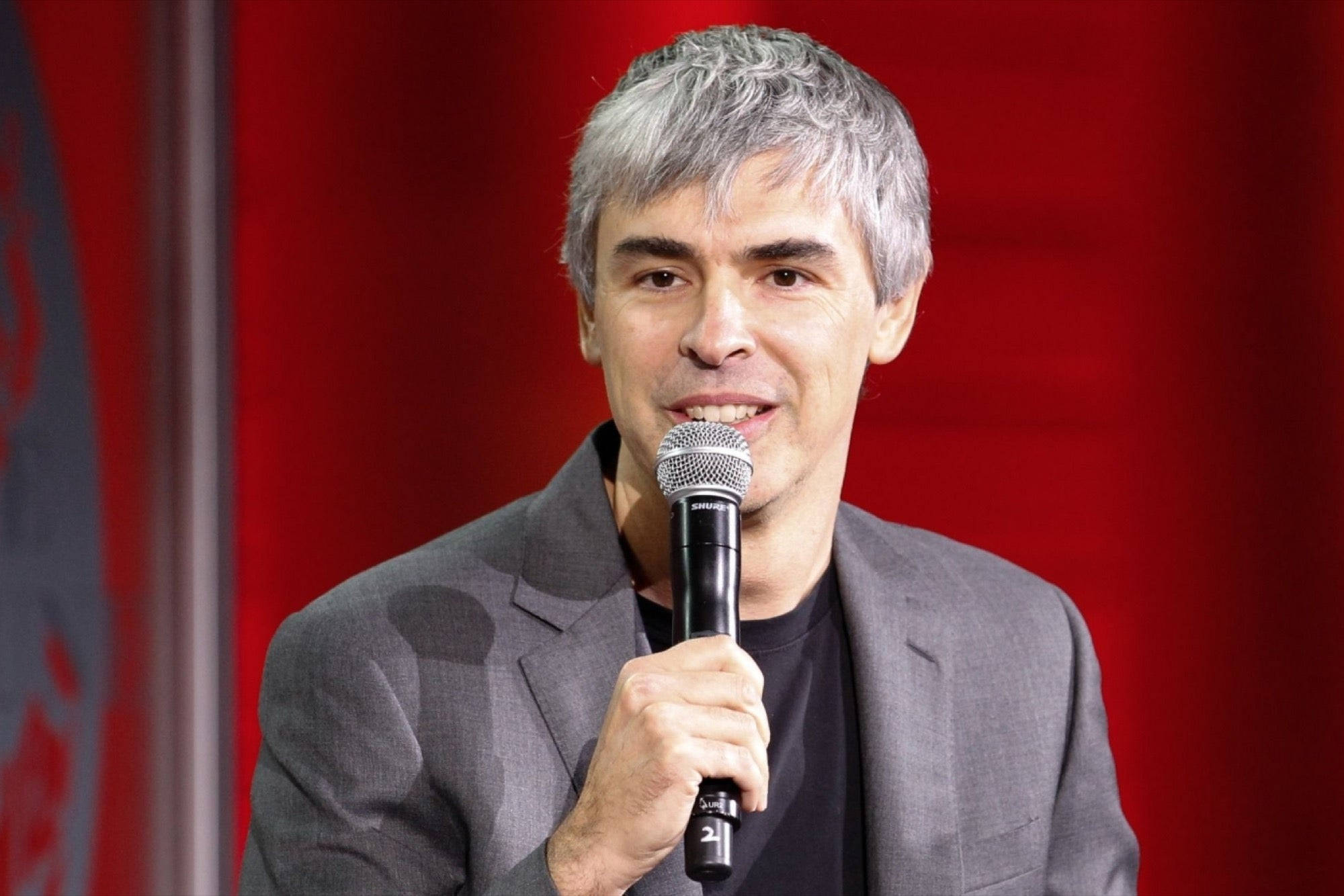 google 桌布 壁紙 — 一位灰髮男士微笑著，手持麥克風，背景是鮮豔的紅色。