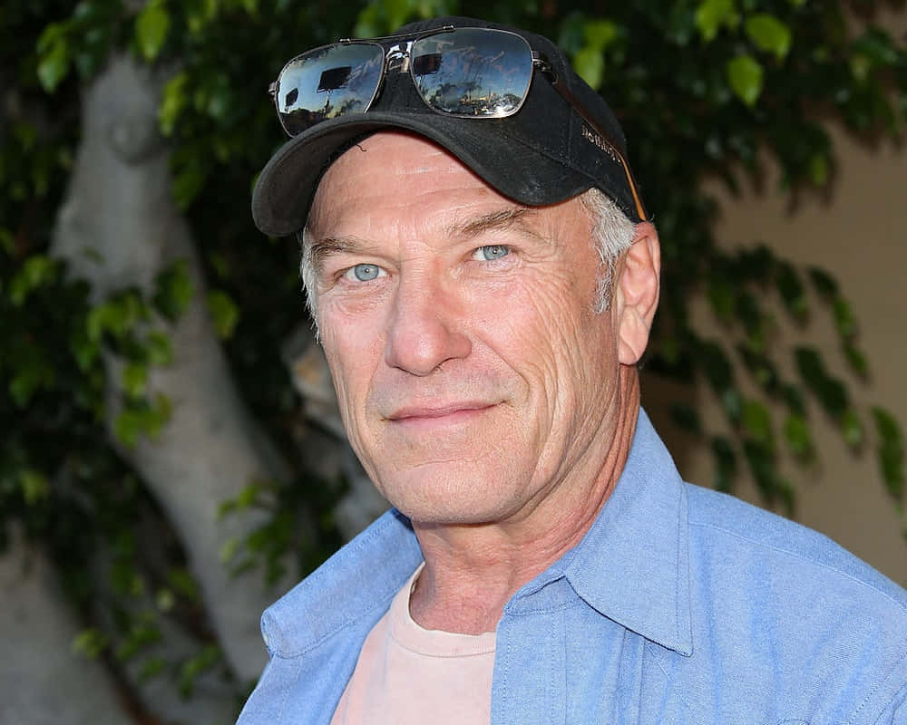Ted Levine 桌布 壁紙 — 一位灰髮男士的特寫肖像，戴著黑色鴨舌帽，太陽眼鏡架在帽上，戶外站立，身後有綠色樹葉。