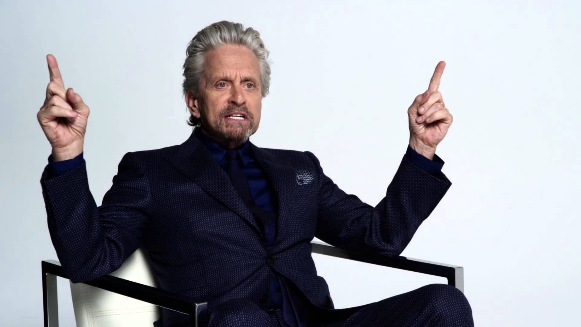 actor 桌布 壁紙 — 一位灰髮男士身穿深色西裝坐在椅子上，雙手食指向上指著純白色背景。