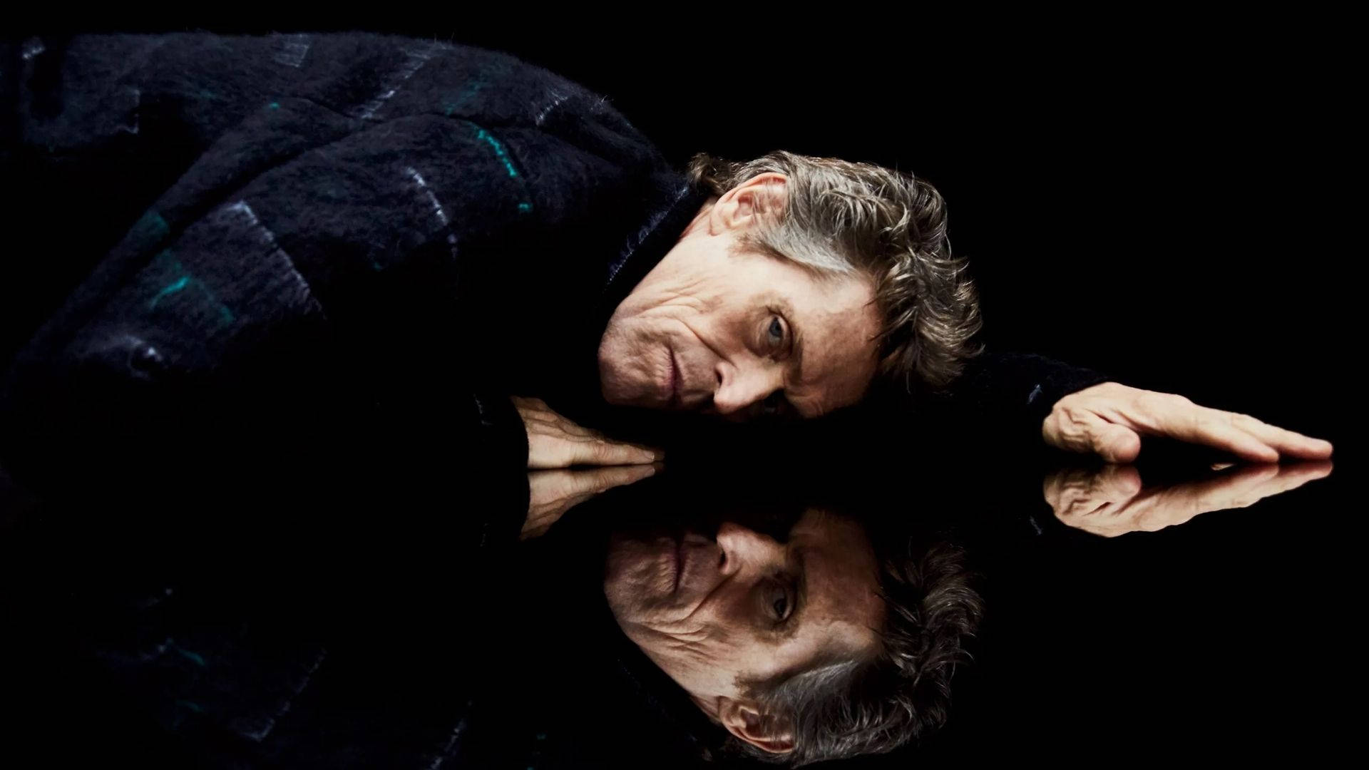 Willem Dafoe 桌布 壁紙 — 一位灰髮男子俯身於反光表面，他的臉和手映照在純黑背景上。