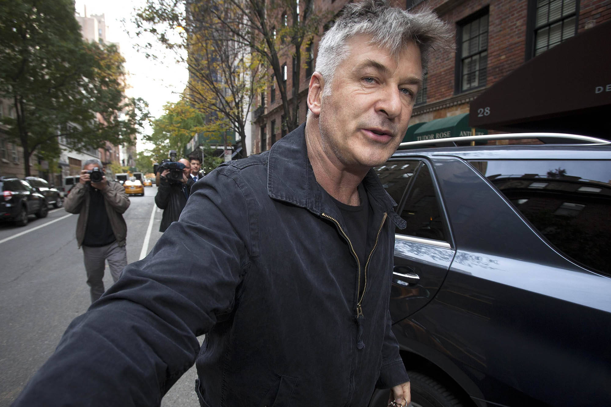 Alec Baldwin 桌布 壁紙 — 一位灰髮男子在晴朗的日子裡沿著城市街道行走，經過攝影師和停泊的汽車。