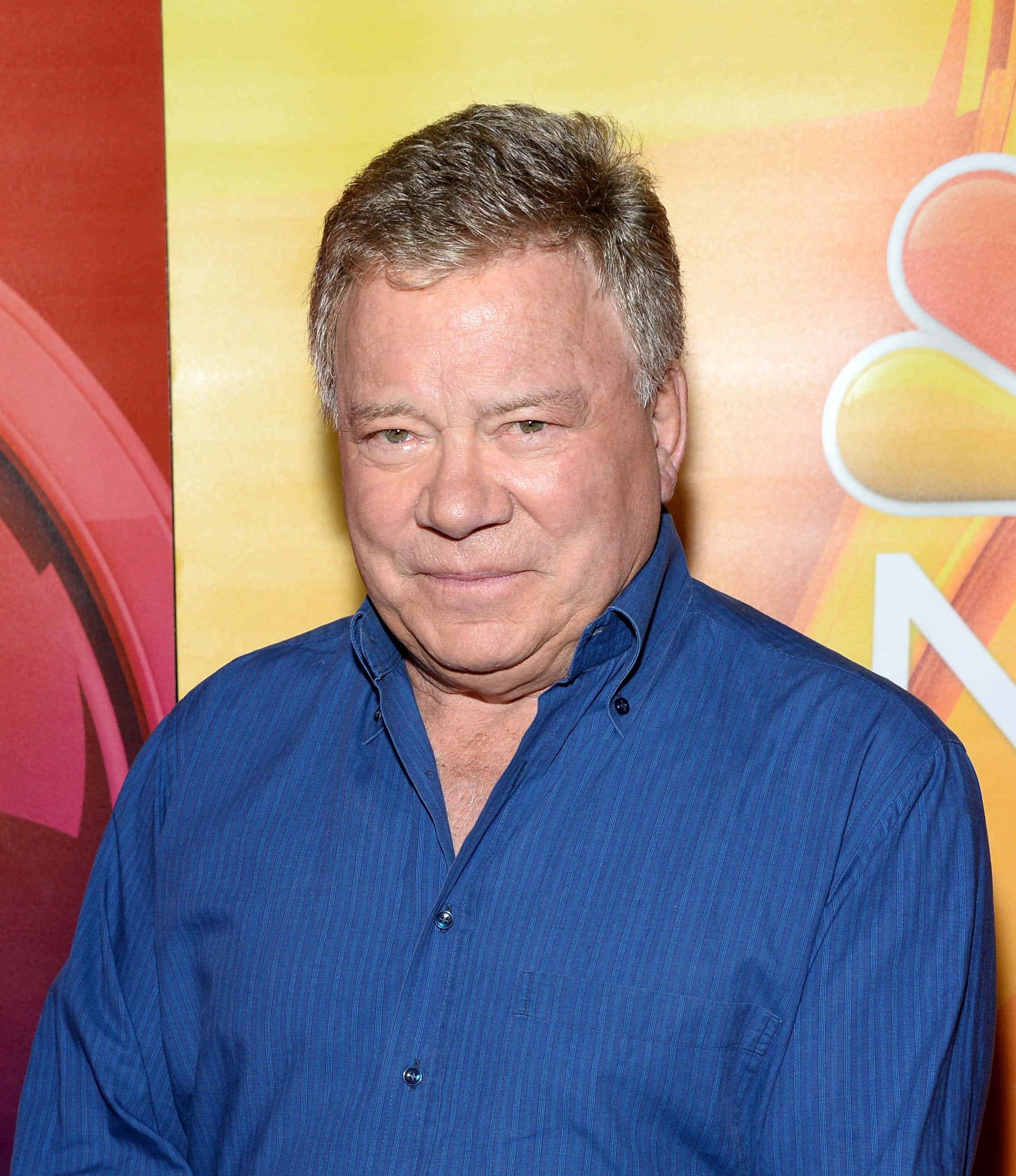 william shatner 桌布 壁紙 — 一位灰髮男子穿著藍色鈕扣襯衫，背景是色彩繽紛的NBC標誌。