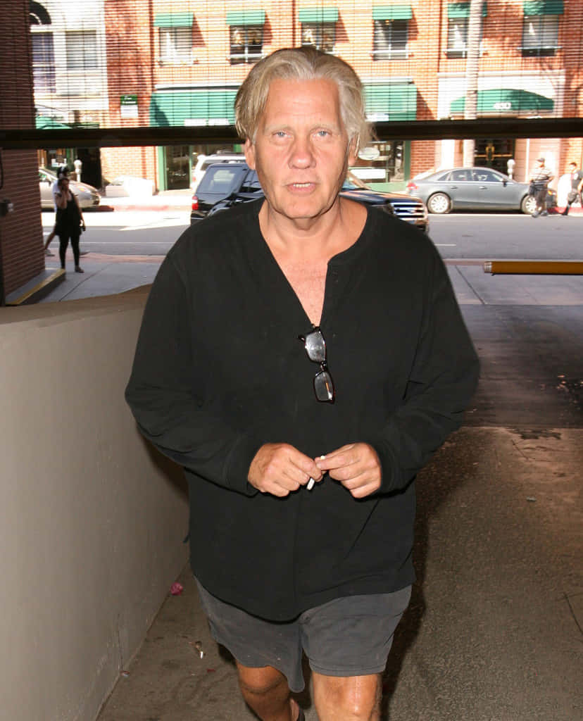 William Forsythe 桌布 壁紙 — 一位灰髮男子穿著黑色襯衫，太陽眼鏡掛在領口，走在城市人行道上。