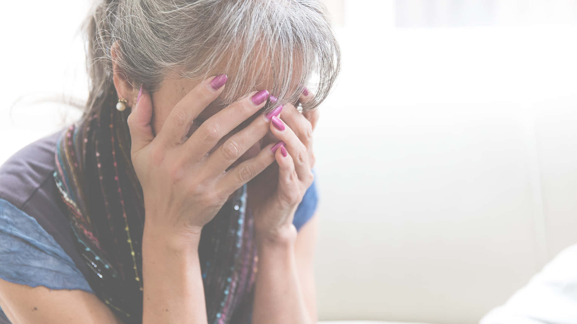 Pain 桌布 壁紙 — 一位灰髮的年長女性在柔和光線的室內環境中用手遮住臉，表現出痛苦。