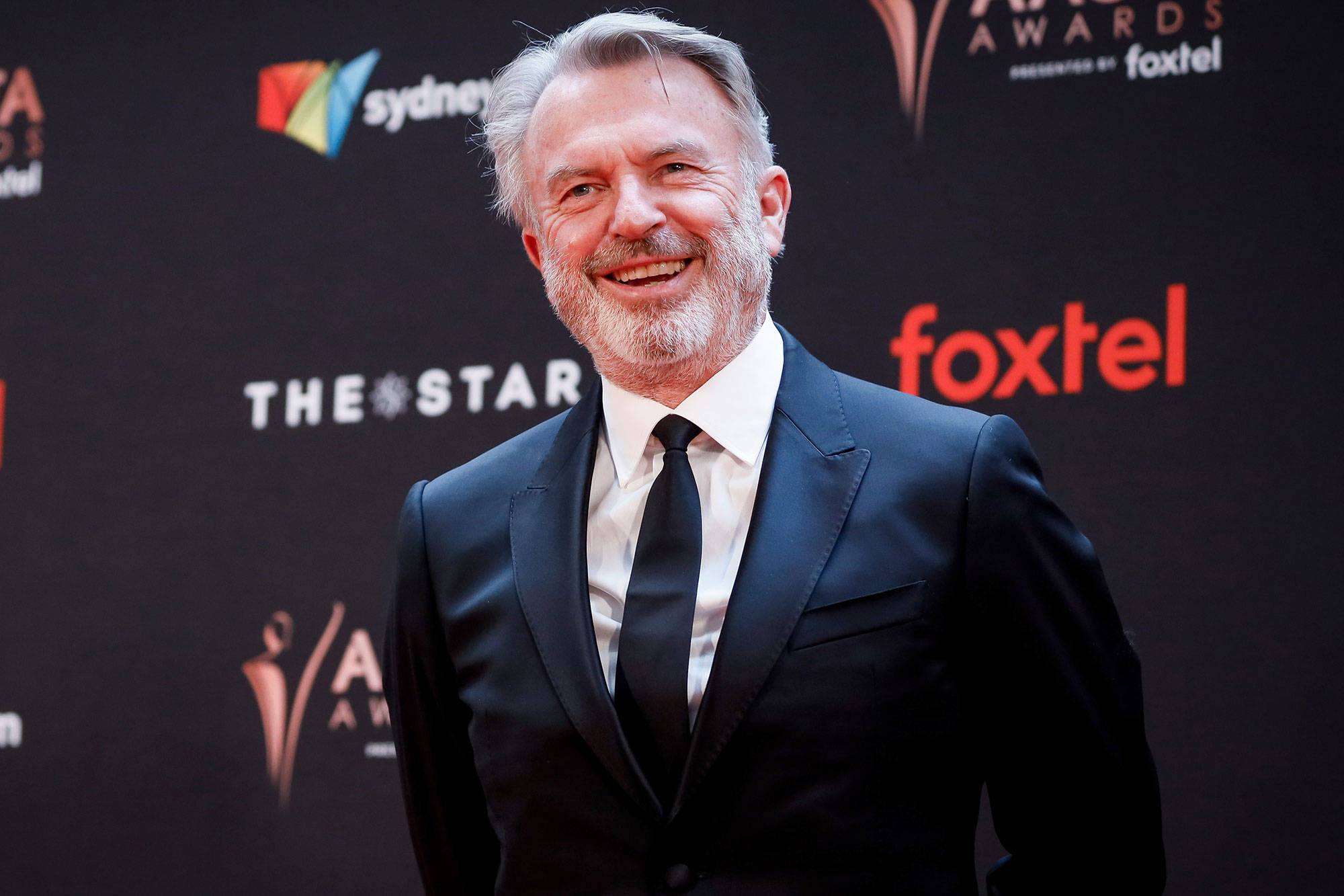 Celebrity 桌布 壁紙 — 一位灰髮鬍鬚、笑容滿面的男士身穿黑色西裝，自信地站在雪梨AACTA獎項的紅地毯上。