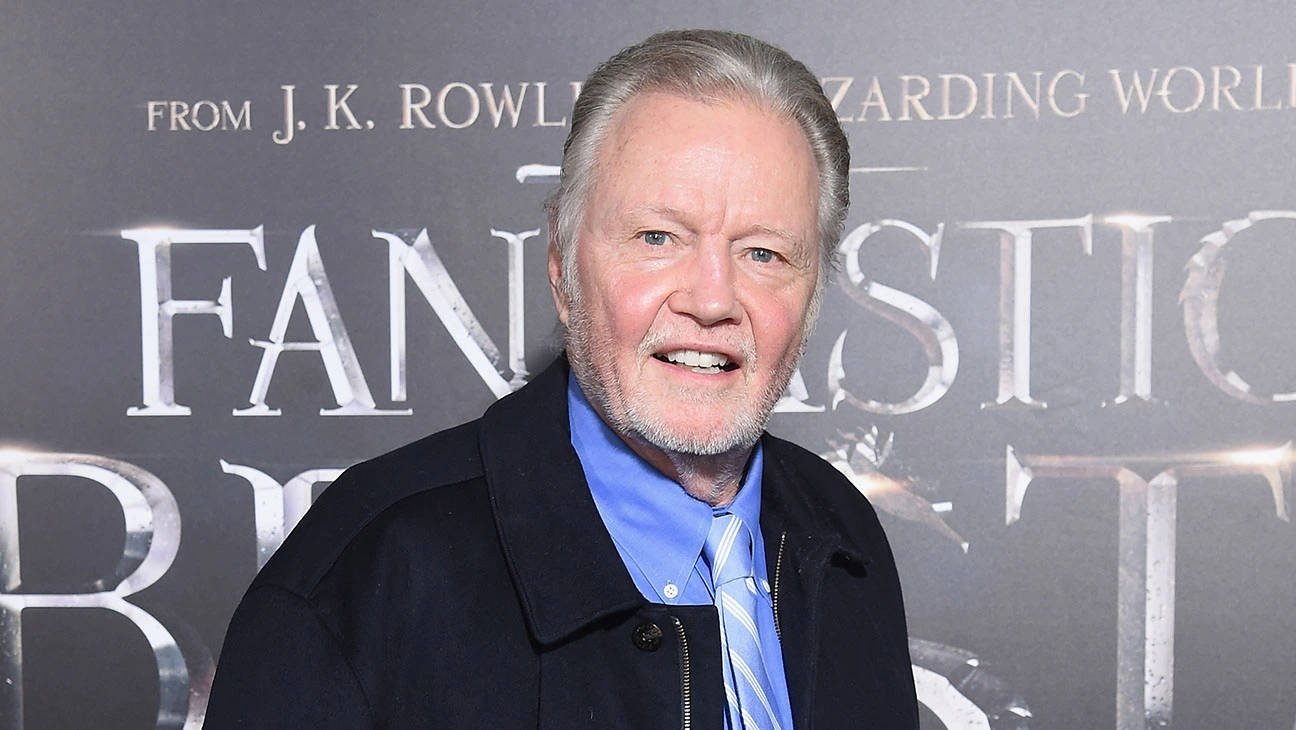 Jon Voight 桌布 壁紙 — 一位灰髮鬍鬚男士穿著深色外套、藍色襯衫和領帶，在《怪獸與牠們的產地》電影背景前對著鏡頭微笑。