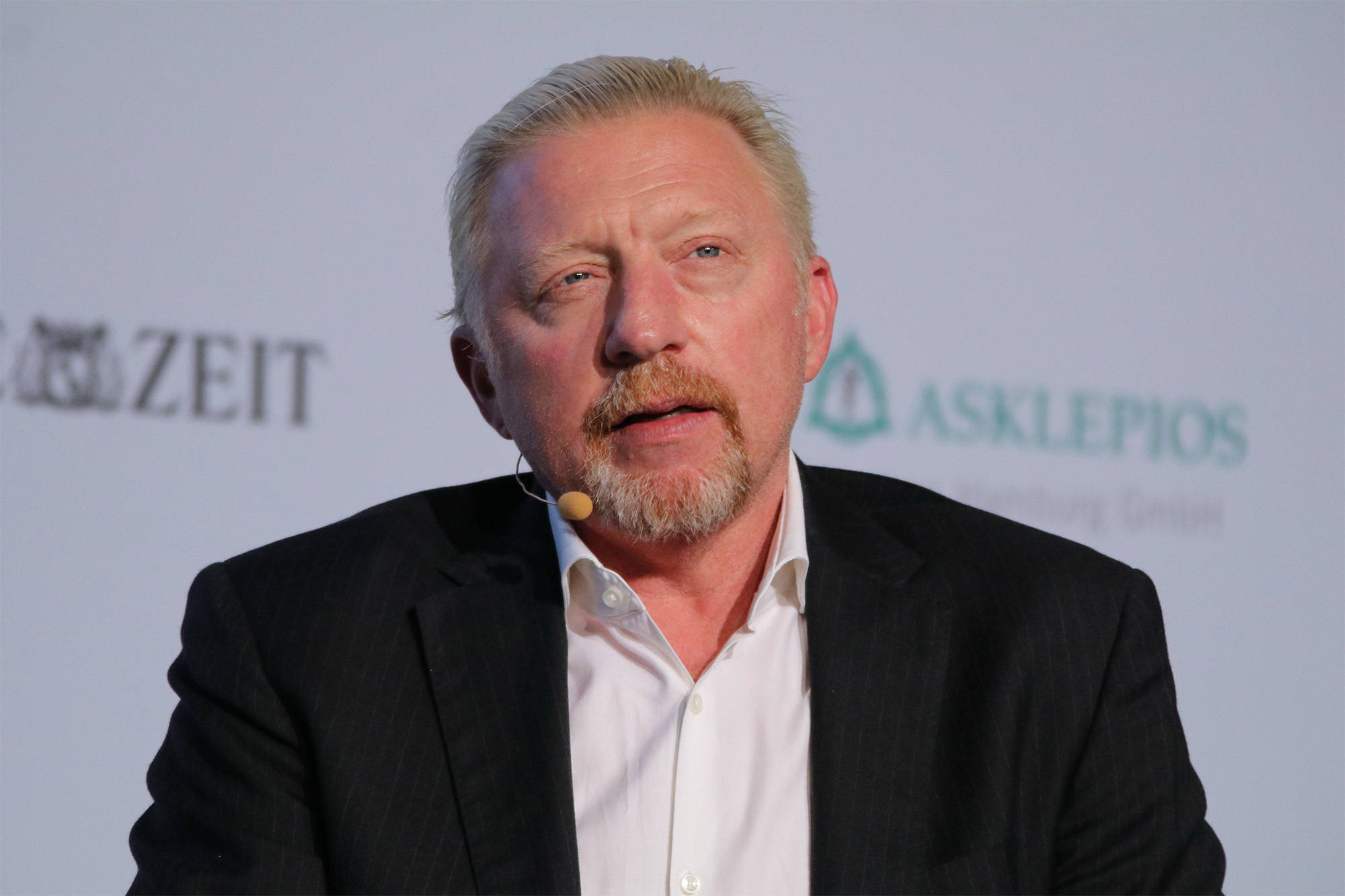 Boris Becker 桌布 壁紙 — 一位灰髮鬍鬚的中年男子在背後有標誌的正式活動中對著麥克風發言。
