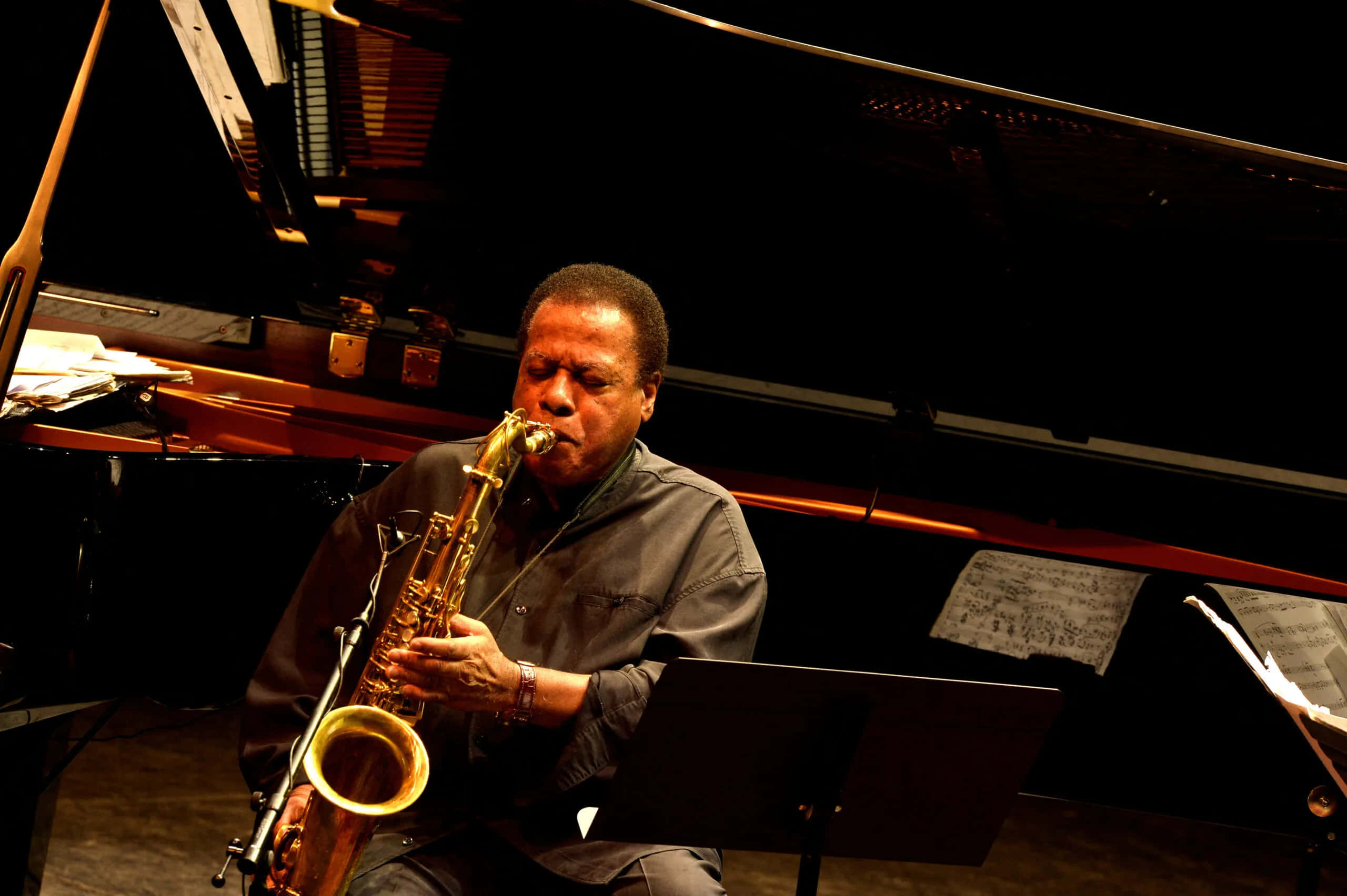 Wayne Shorter 桌布 壁紙 — 一位爵士樂手在溫暖的舞台燈光下，於三角鋼琴旁熱情地吹奏金色薩克斯風。