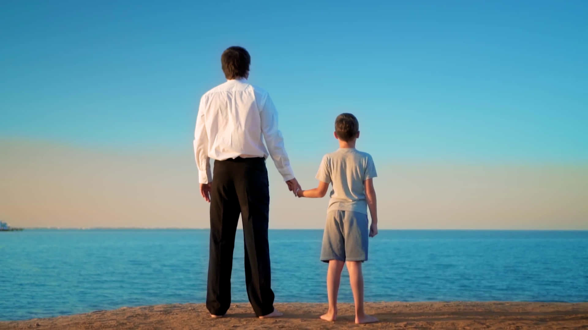 Father And Son 桌布 壁紙 — 一位父親和兒子手牽手，在晴朗的藍天下，日落時分凝視著平靜的海洋。