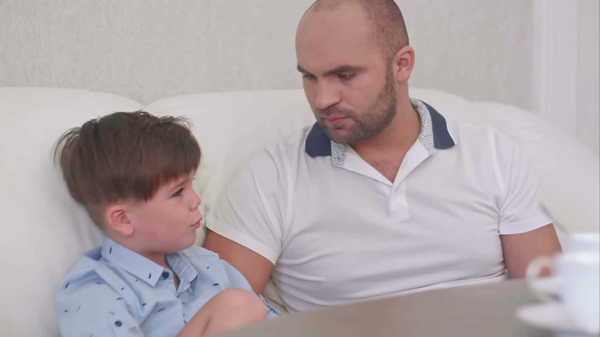 Father And Son 桌布 壁紙 — 一位父親和年幼的兒子緊密地坐在白色沙發上，分享一個安靜親密的室內時刻。