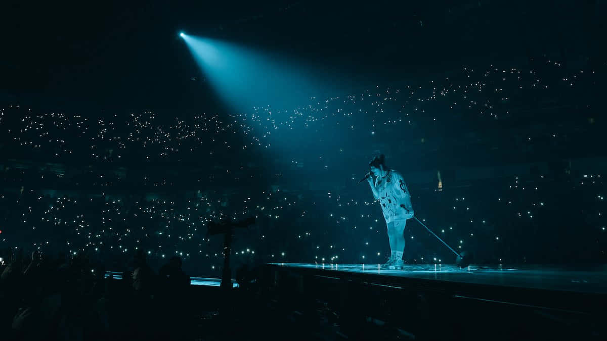 Billie Eilish Laptop 桌布 壁紙 — 一位獨唱表演者站在黑暗的舞台上，沐浴在單一藍色聚光燈下，被觀眾手機燈光的海洋包圍。