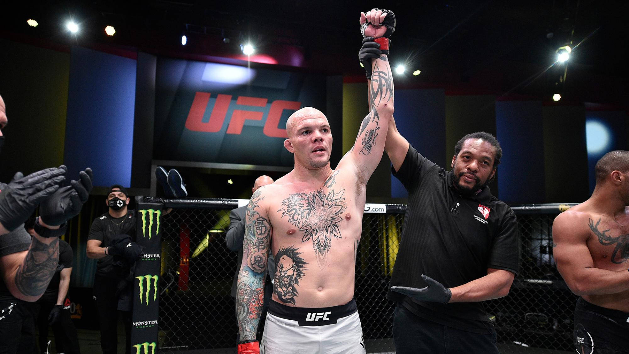Champion 桌布 壁紙 — 一位獲勝的UFC拳手，身上有複雜的紋身，在明亮的場館燈光下被教練在八角籠中舉起。