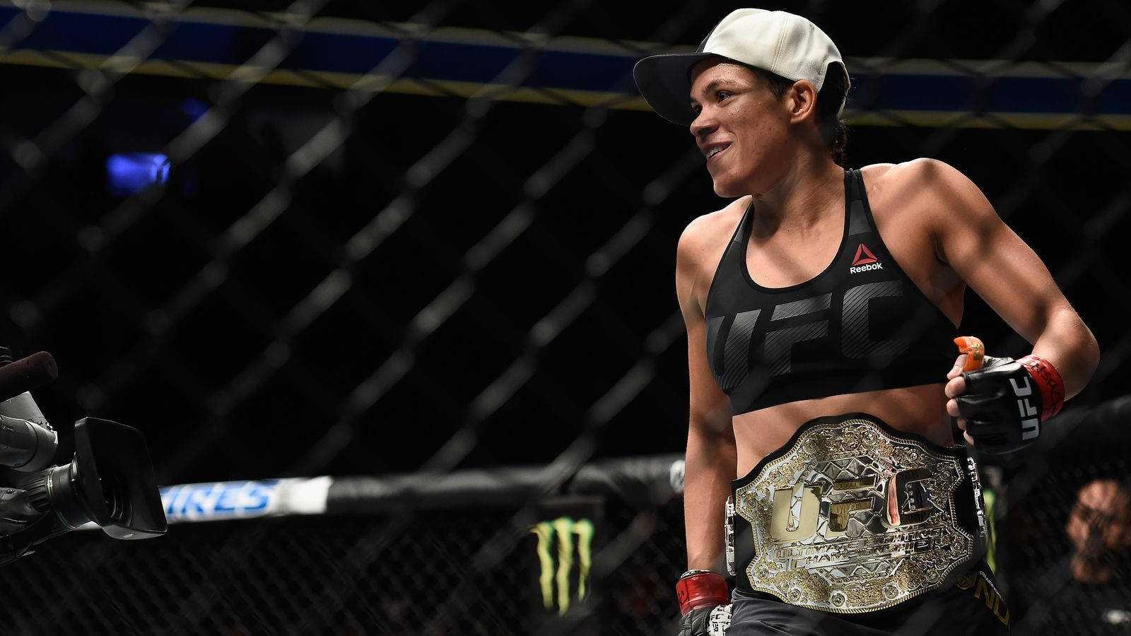 Champion 桌布 壁紙 — 一位獲勝的女性MMA拳手在八角籠內自豪地微笑，佩戴著冠軍腰帶和品牌裝備。