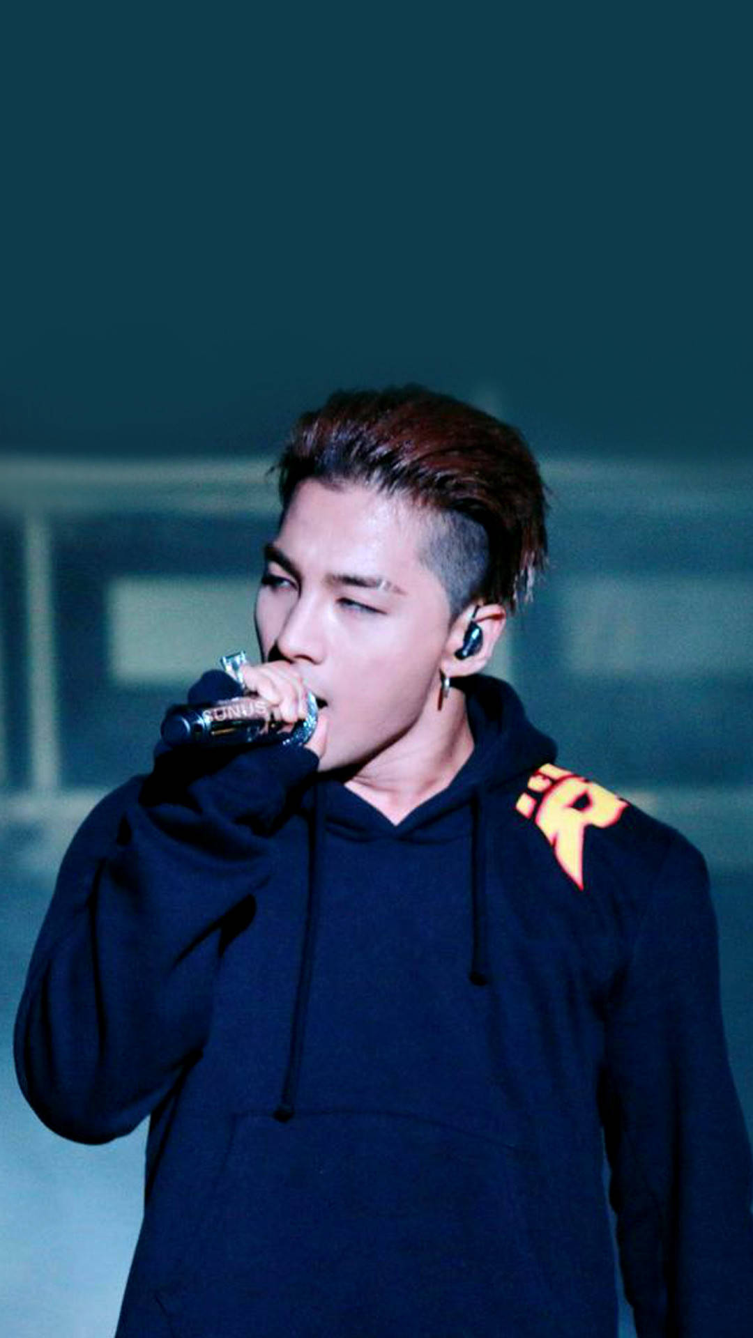 Bigbang 桌布 壁紙 — 一位男性表演者在冷藍色舞台燈光下對著麥克風唱歌，身穿帶有橙色字母的深色連帽衫。