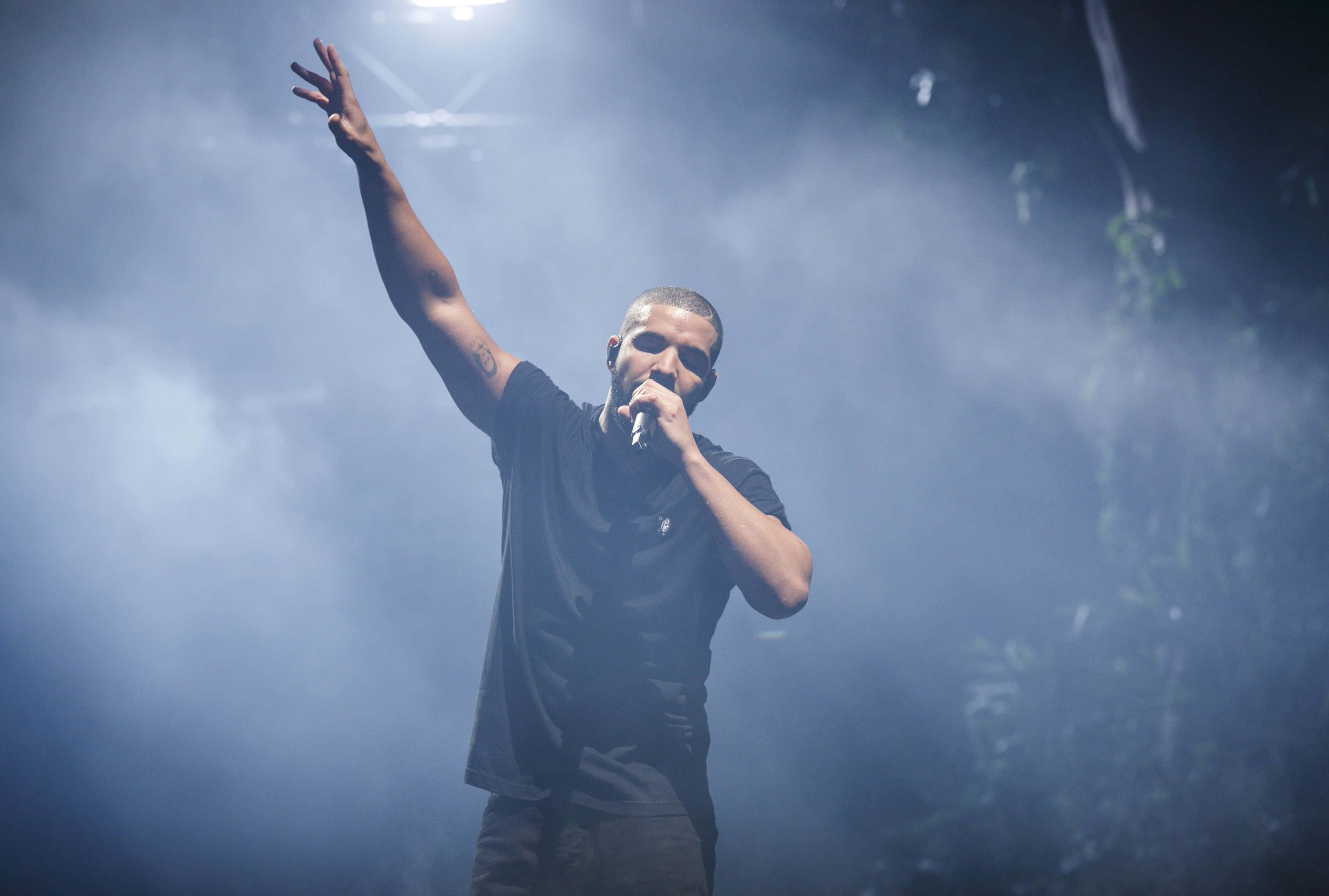 Drake 桌布 壁紙 — 一位男性表演者在煙霧瀰漫的舞台上，在戲劇性的藍色燈光下，舉起一隻手臂對著麥克風唱歌。