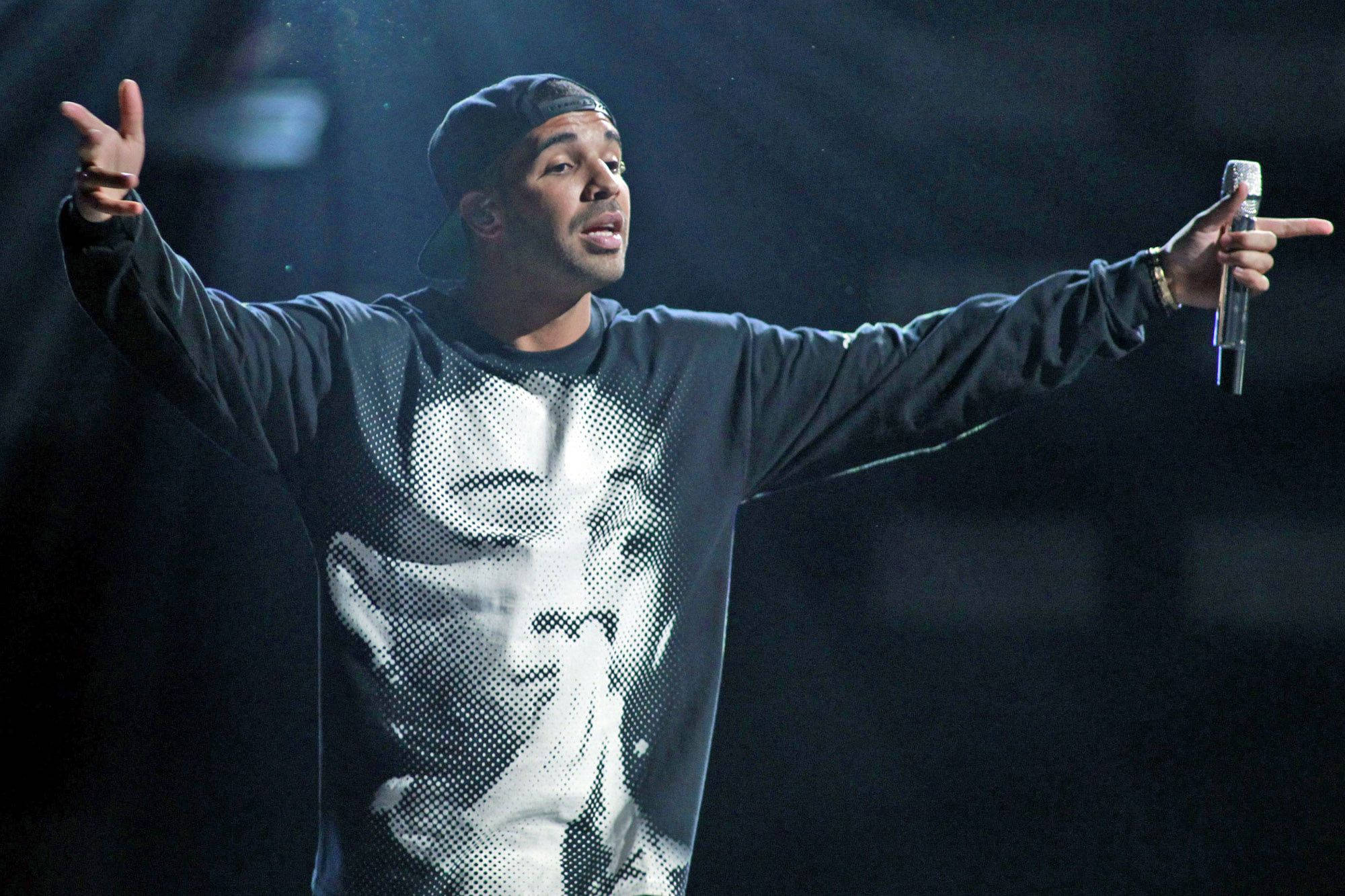 Drake 桌布 壁紙 — 一位男性表演者戴著黑色帽子，穿著圖案衛衣，在黑暗的舞台上張開雙臂，握著麥克風熱情地唱歌。