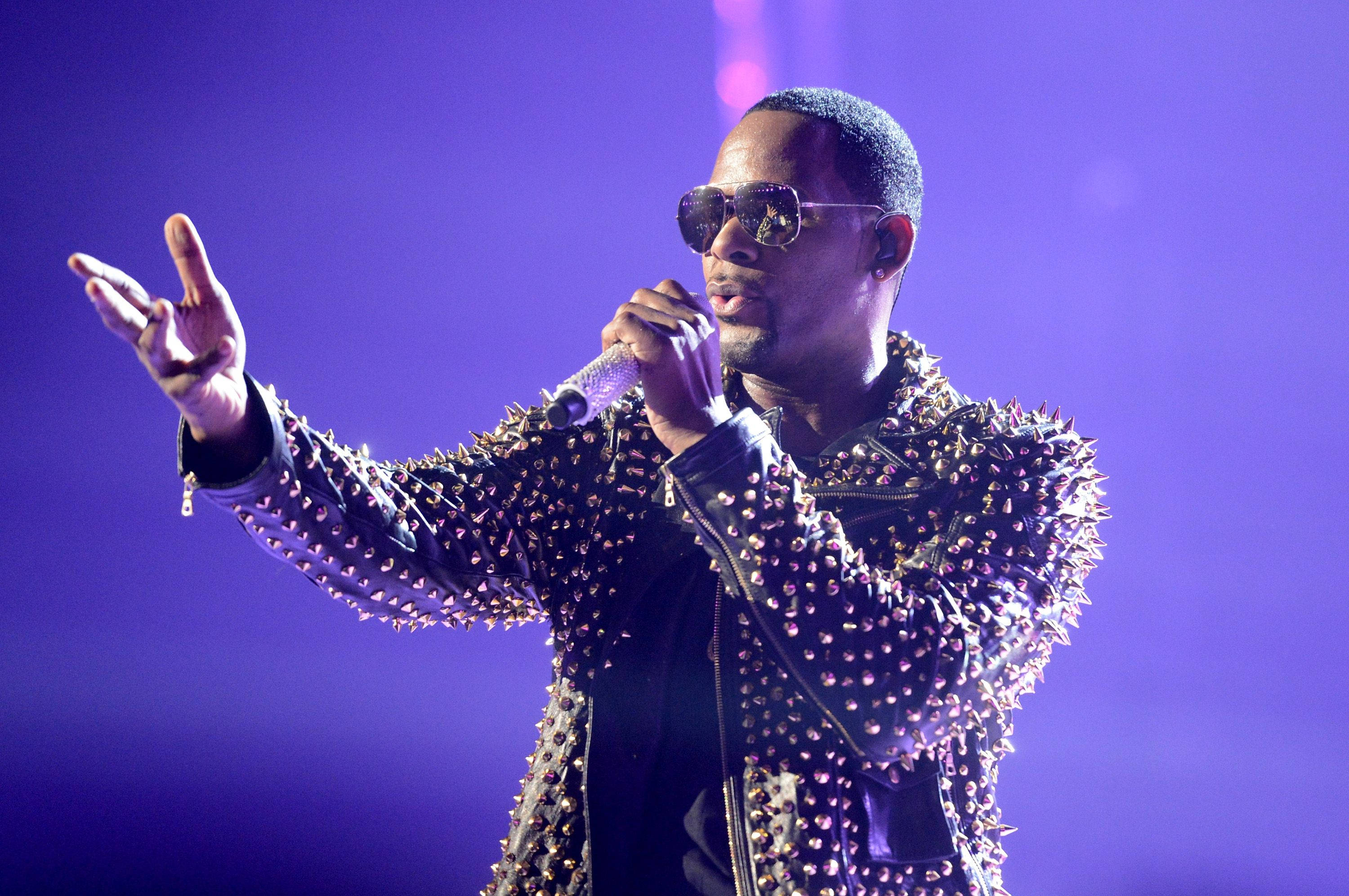 r kelly 桌布 壁紙 — 一位男性表演者穿著鑲有鉚釘的皮夾克和太陽眼鏡，在紫色舞台燈光下對著麥克風唱歌。