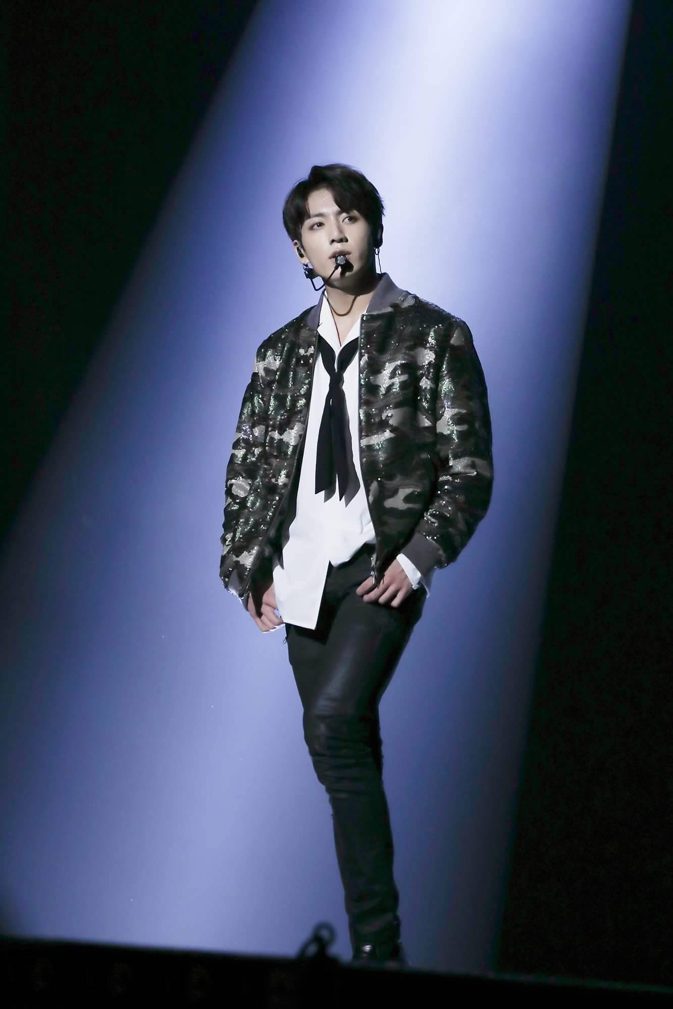 BTS 桌布 壁紙 — 一位男性表演者身穿迷彩外套和黑色褲子，在黑暗舞台上的聚光燈下自信地站立。