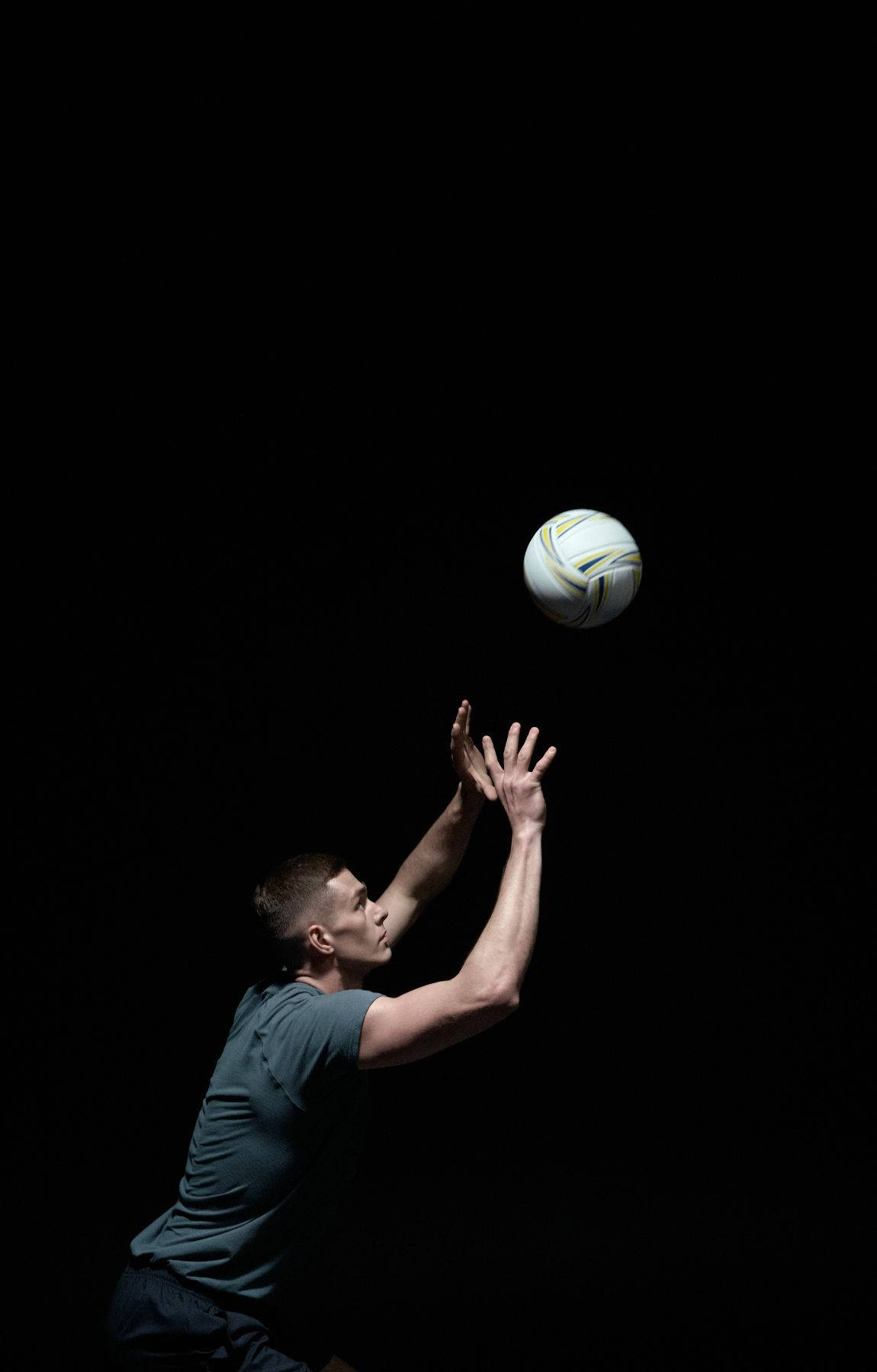 Volleyball Aesthetic 桌布 壁紙 — 一位男性運動員在昏暗的攝影棚中向上伸手，在純黑色背景前接住一個白色排球。