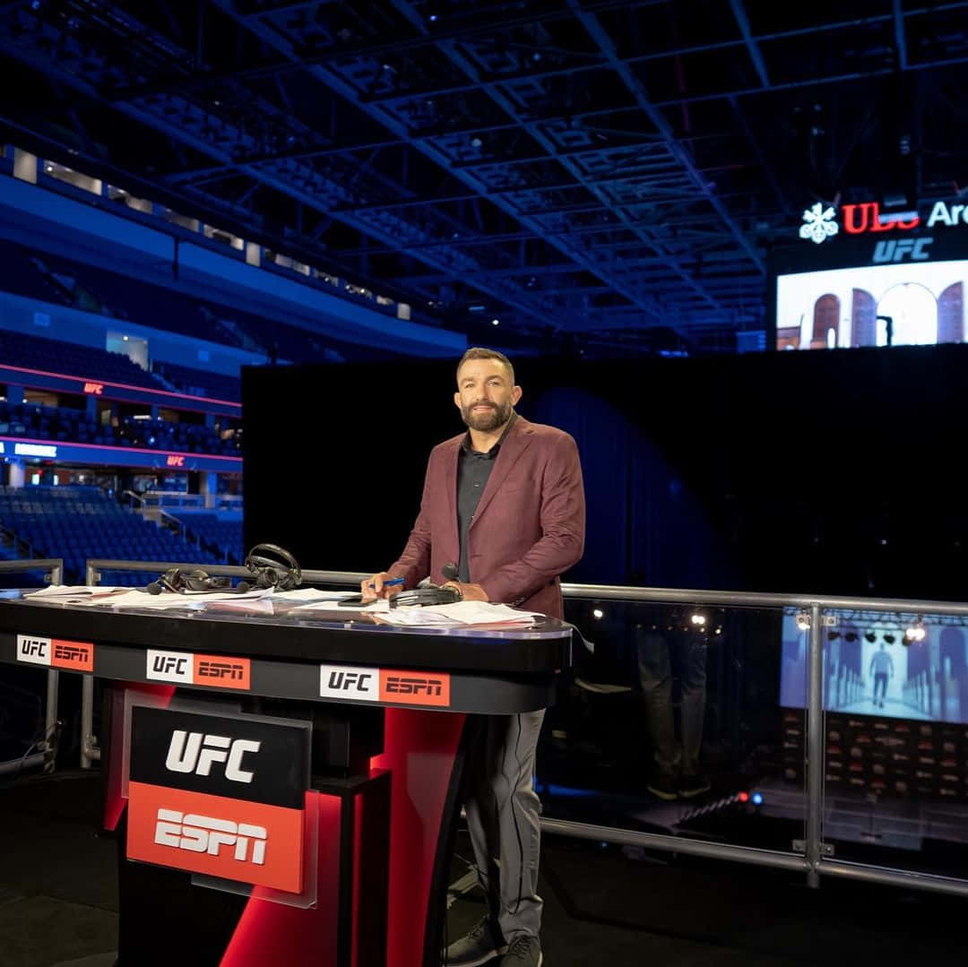 運動 桌布 壁紙 — 一位男性體育評論員站在UFC ESPN轉播台前，身處燈光昏暗、藍色燈光且座位空無一人的競技場。