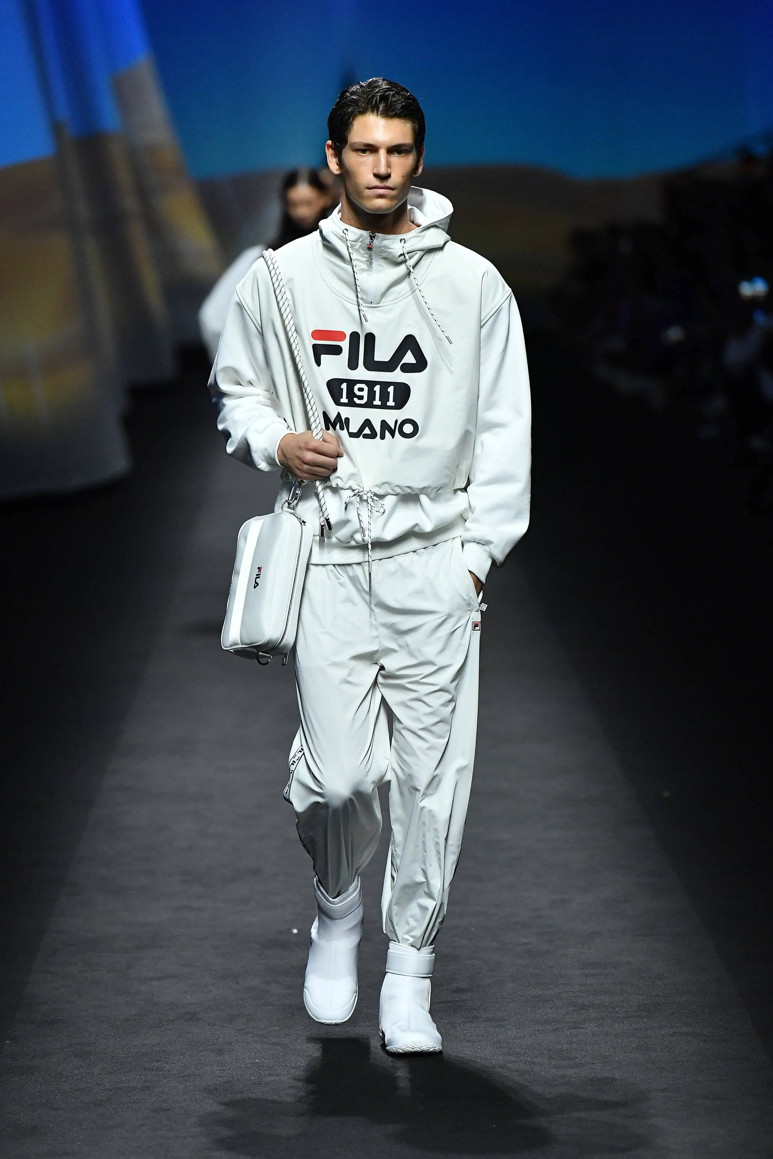 Fila 桌布 壁紙 — 一位男模特兒在戲劇性的舞台燈光下，穿著全白色的Fila運動套裝和相配的包包，自信地走在天橋上。