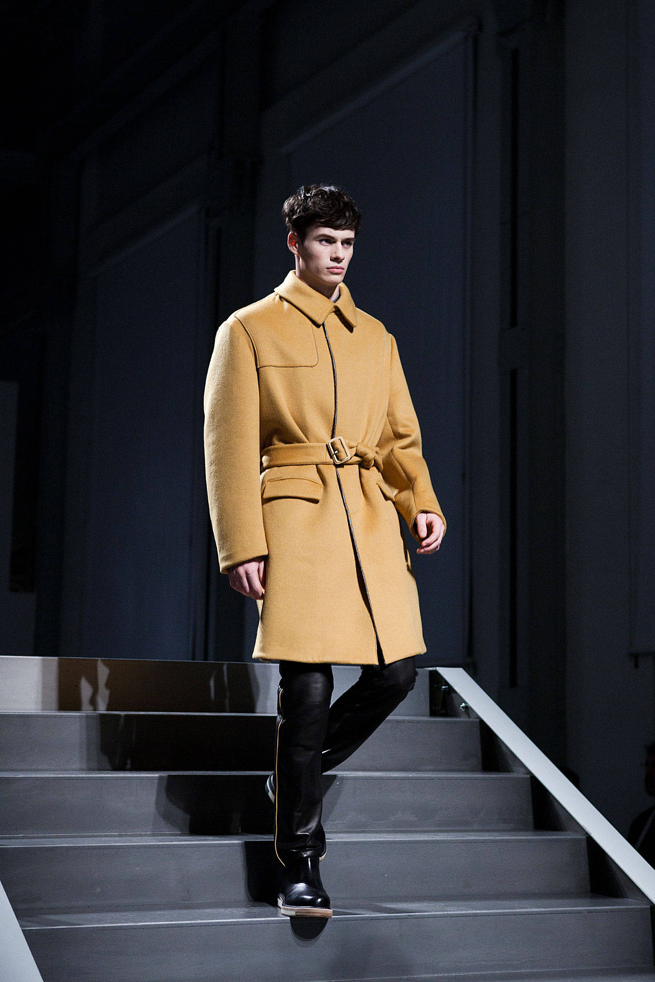 Fendi 桌布 壁紙 — 一位男模特兒穿著芥末黃色束帶外套和黑色靴子，在戲劇性的伸展台燈光下自信地走下現代樓梯。